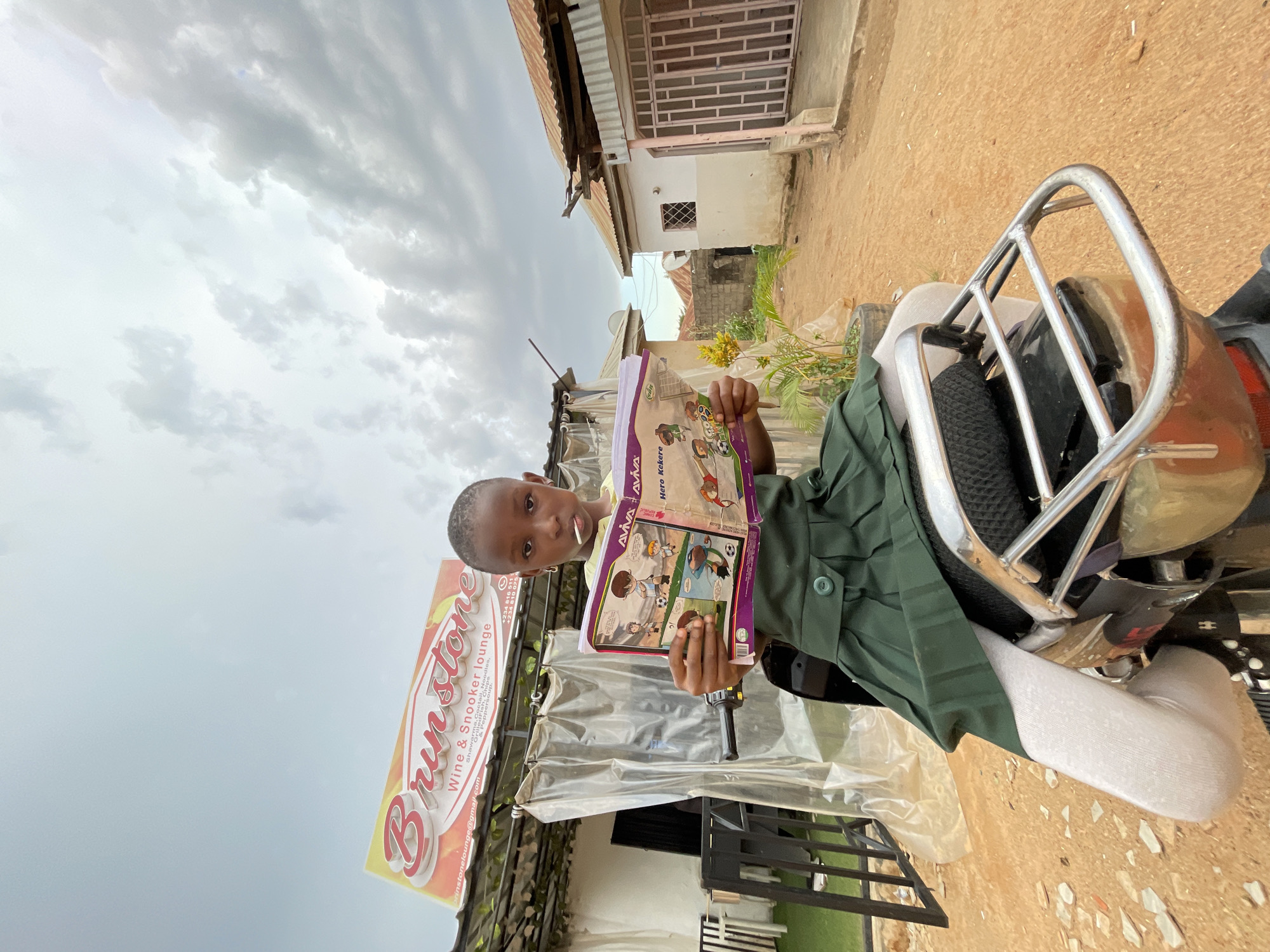 A young, African child sits on a motorcycle holding a purple children's book with illustrated characters titled "Hero Kekere," by Aviva. The child wears a green, button-down dress, and light-colored socks. The background shows a street scene with a building displaying a sign for "Brinstone Wine & Snooker Lounge" visible, indicating the location is likely in Jos, Nigeria. The sky is overcast, and surrounding structures suggest a developing urban environment.