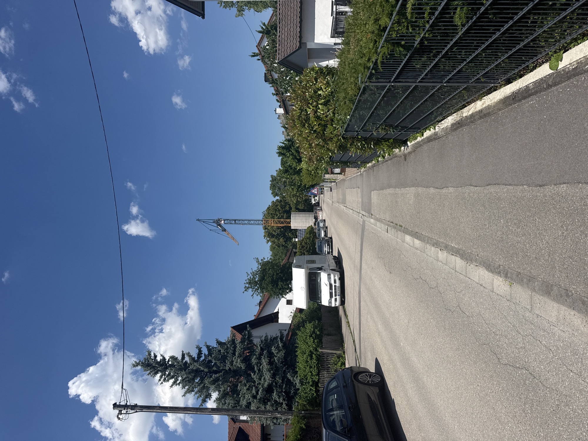 The image depicts a street scene in Höhenkirchen-Siegertsbrunn, Germany, on a sunny day with a clear blue sky and scattered white clouds. A residential street is visible, with houses and dense green hedges lining both sides. A black car is parked on the left side of the road, partially in frame. Further down the street, a white camper van is parked, followed by a line of other cars. A large construction crane is prominent in the background, indicating ongoing development. A sidewalk runs along the right side of the street, separated from the road by a curb. A dark metal fence borders the sidewalk, with lush green bushes behind it. A wooden utility pole with electrical wires is situated on the left side of the road, near a tall evergreen tree.