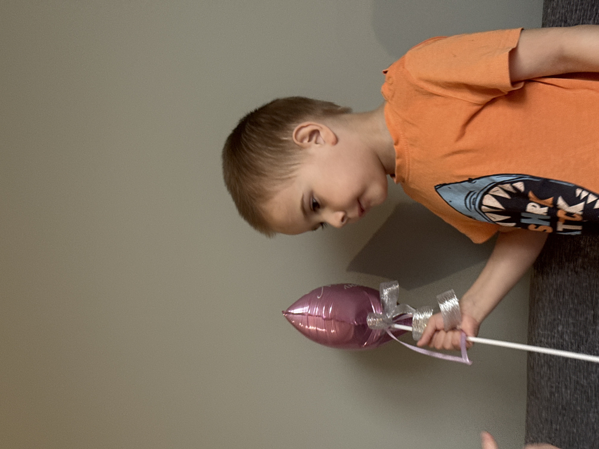 A young boy in an orange t-shirt with a shark graphic is looking down at a small, purple, heart-shaped balloon attached to a white stick with a silver ribbon. The setting appears to be indoors with a light gray wall in the background. The location is not discernible from the visual information provided, and there is no indication of Chaikovsky, Russia.