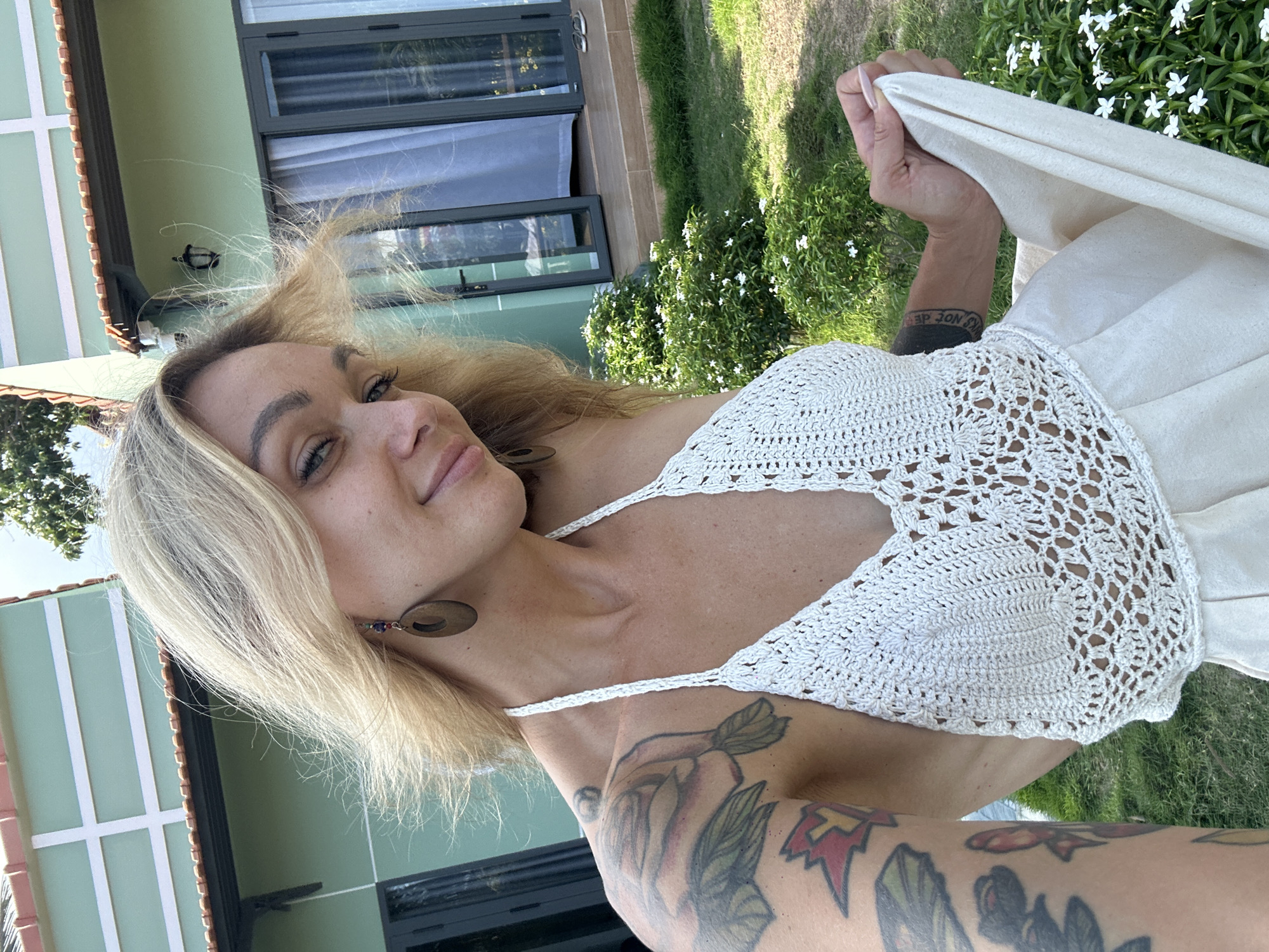 A woman is posing for a selfie-style shot outdoors. She is wearing a white crocheted top and a light-colored skirt or dress, and she's holding a piece of white fabric, likely part of the outfit. The location appears to be in Mũi Né, Vietnam, given the presence of tropical plants and the style of architecture visible in the background. The woman has tattoos on her arms and is wearing earrings. The background includes a building with light green walls and dark-framed windows, as well as a grassy area with white flowers. The camera angle is slightly tilted.