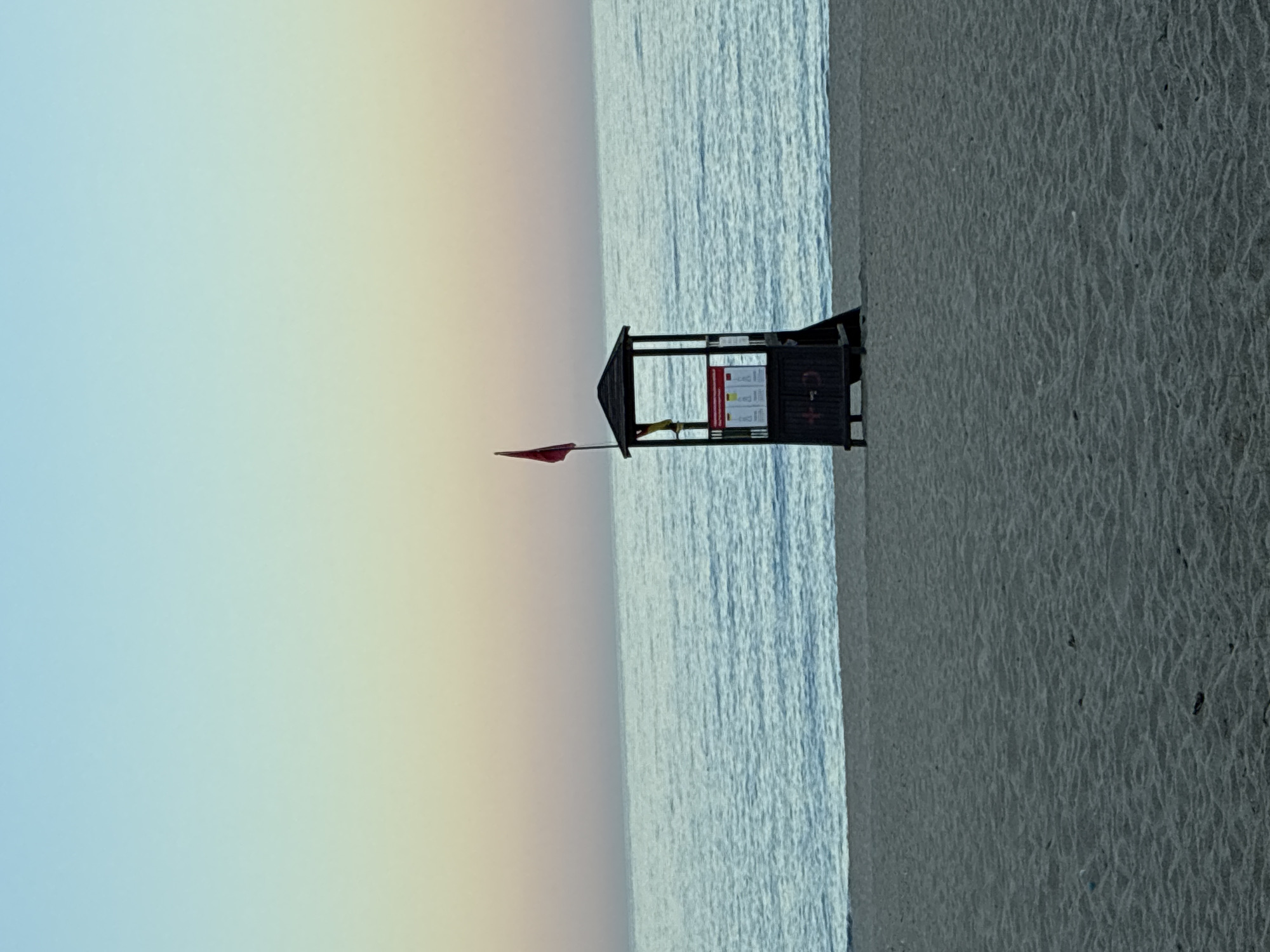 A lifeguard tower stands on a sandy beach in Alanya, Turkey. The tower is dark brown with a dark roof and a red flag flying from its top. A yellow flag is also visible hanging from the tower. The tower has a sign with red lettering and some smaller documents displayed on its front. The sand is mostly flat with some ripples. The sea is a calm, light blue, and the sky transitions from a pale blue at the top to a soft pink and yellow near the horizon. The scene appears to be during sunrise or sunset. There are no people visible in the image.