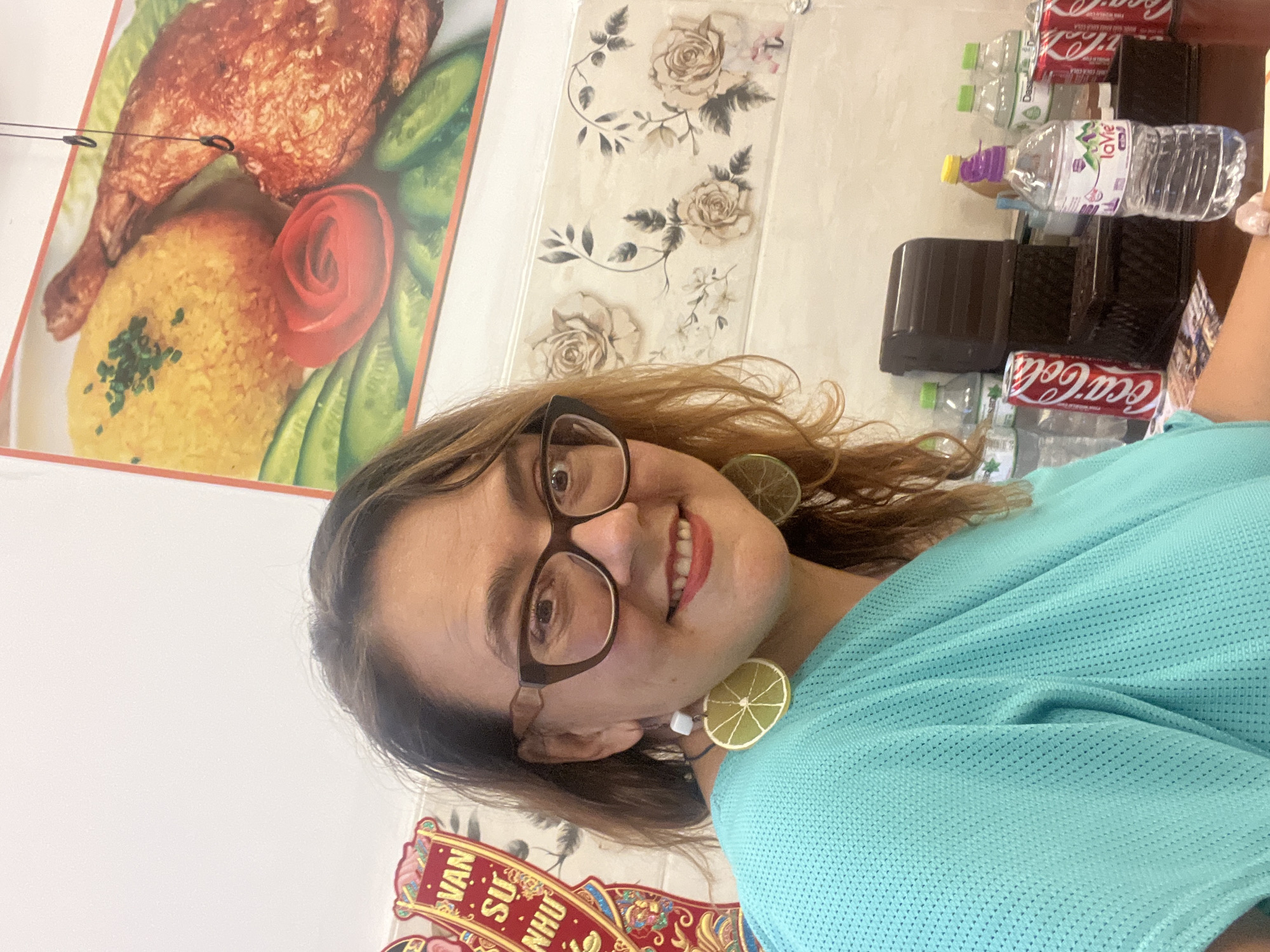 A medium shot shows a woman with brown hair and glasses in a turquoise shirt, smiling at the camera. She is wearing large, circular earrings that resemble lime slices. Behind her, a menu board displays an image of chicken and rice, and a wall is decorated with a floral pattern and red and gold banners. On a table in front of her are several bottles of water, two cans of Coca-Cola, and a dark brown box. The setting appears to be a casual eatery in Nha Trang Ward, Vietnam.