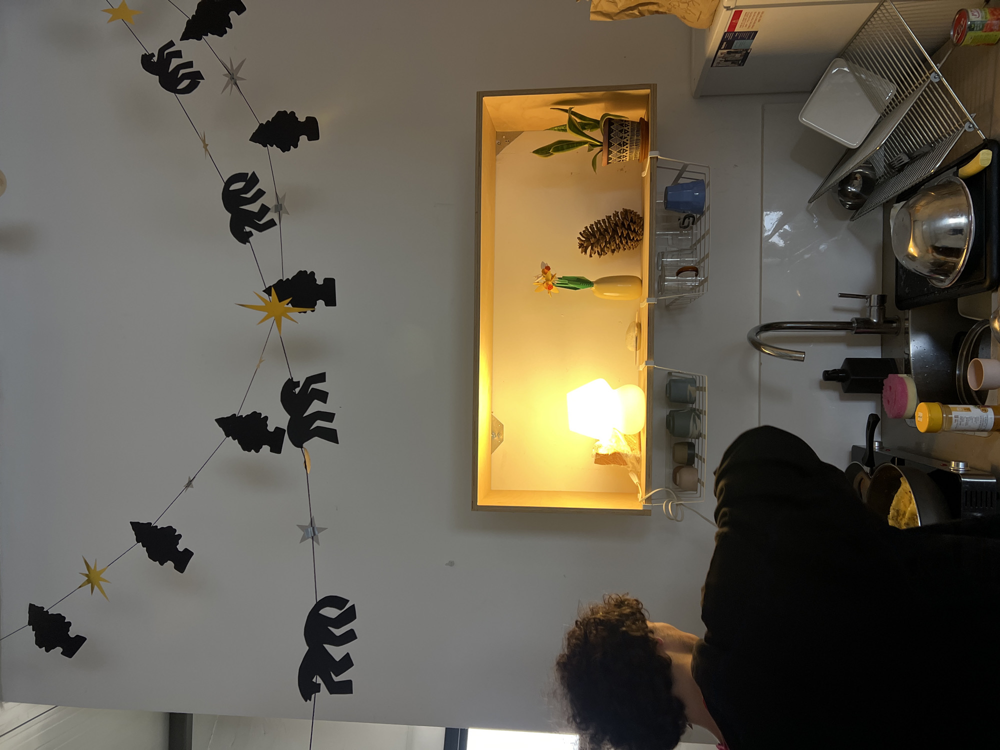 The image depicts an indoor kitchen space in a residential dwelling located in Tel-Aviv, Israel.
A person with dark, curly hair, viewed from behind, wearing a dark long-sleeved garment, is actively cooking in the lower-left quadrant of the frame. The individual is positioned over a stovetop where food, appearing yellowish, is contained within a dark pan. To the right of the cooking area, a yellow spice container and a pink scrubbing sponge are visible on the countertop.
Above the countertop, a stainless steel sink features a modern chrome faucet and a black pump dispenser. Adjacent to the sink on the right, a dish drying rack holds a large silver mixing bowl, a white square plate, and other utensils. A small yellow object is near the bowl.
Above the sink area, a recessed wooden shelf unit is brightly illuminated by an internal light source, casting a warm yellow glow. On this shelf, from left to right, are a white mushroom-shaped table lamp, a yellow vase holding an artificial daffodil-like flower, a large pinecone, and a potted plant with green leaves in a patterned terracotta pot. A small white circular object rests to the left of the vase. Attached beneath this shelf are wire racks holding several drinking glasses and a blue mug.
The upper wall area is adorned with a string of black paper cutouts depicting stylized trees and horses, interspersed with yellow and silver star cutouts, forming two upward-curving garlands. A portion of a white refrigerator is visible on the far right. A brown paper bag and a red can are also partially visible on the right side of the counter.