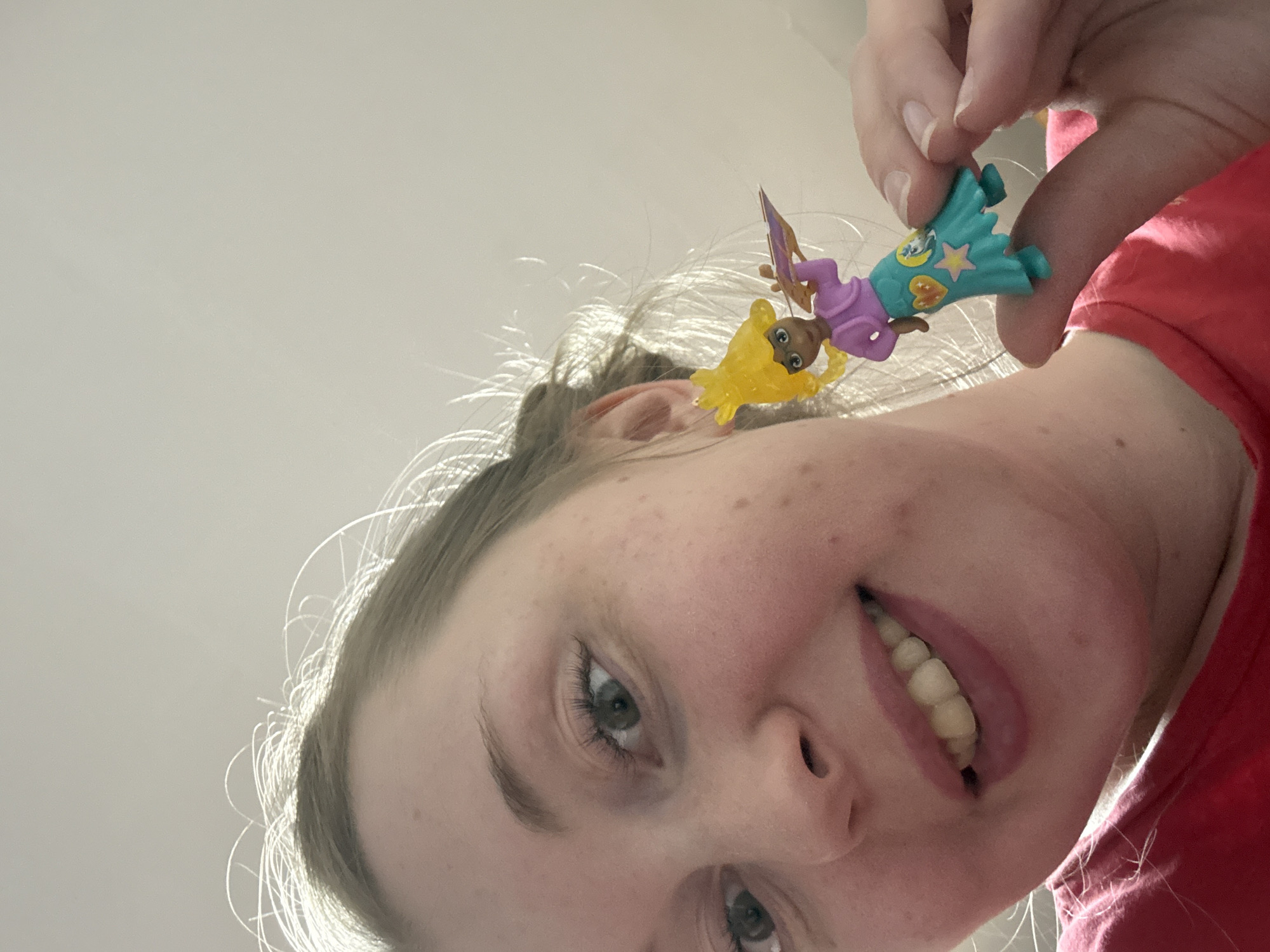 A person is holding a small, colorful toy figure in front of their face. The figure appears to be a character, possibly from a children's show, with a yellow body, purple top, and turquoise skirt. The person is smiling. The location of the image is unknown.