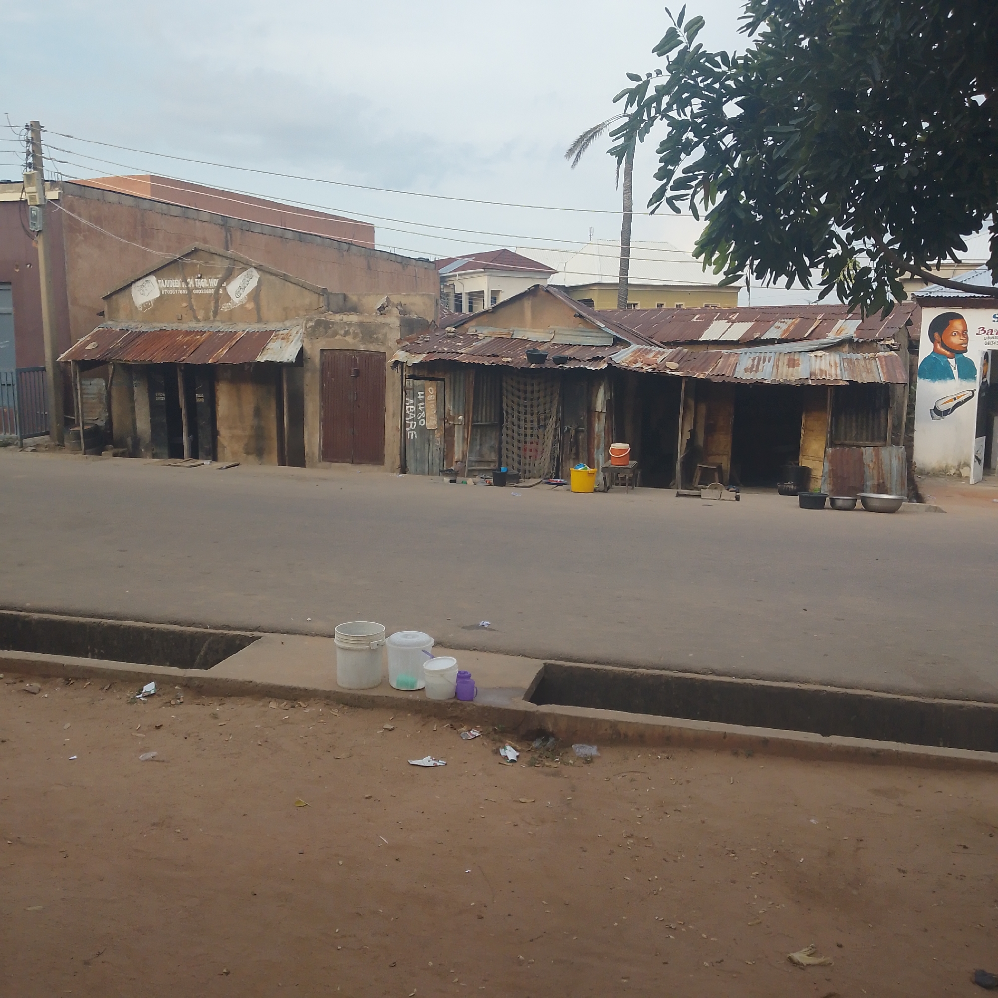 This is a ground-level, eye-level shot of a street scene in Arawa, Nigeria. The scene depicts a row of small, weathered shops with corrugated metal roofs. The shops are built close together along the side of a paved road. To the left, a larger, older building with a faded facade stands behind the shops. A utility pole with wires is visible near this building.
In front of the shops, the road is mostly empty, showing signs of wear and tear. Along the edge of the road, there's a drainage ditch. On the dirt shoulder beside the ditch, several white plastic buckets, a smaller white bucket, and a purple container are placed. Scattered debris, including bits of paper and plastic, litters the ground.
One of the shops has a painted mural on its facade depicting a man's head and shoulders, along with a drawing of an electric shaver and text that appears to be a business name, possibly "BARBER." Other shops have open doorways revealing dark interiors. Various objects, including buckets and metal basins, are placed outside some of the establishments. A tall palm tree and the leaves of a larger tree are visible in the background, suggesting a somewhat tropical or sub-tropical environment. The sky is overcast with a pale, diffused light. The overall impression is one of everyday urban life in a busy commercial area.