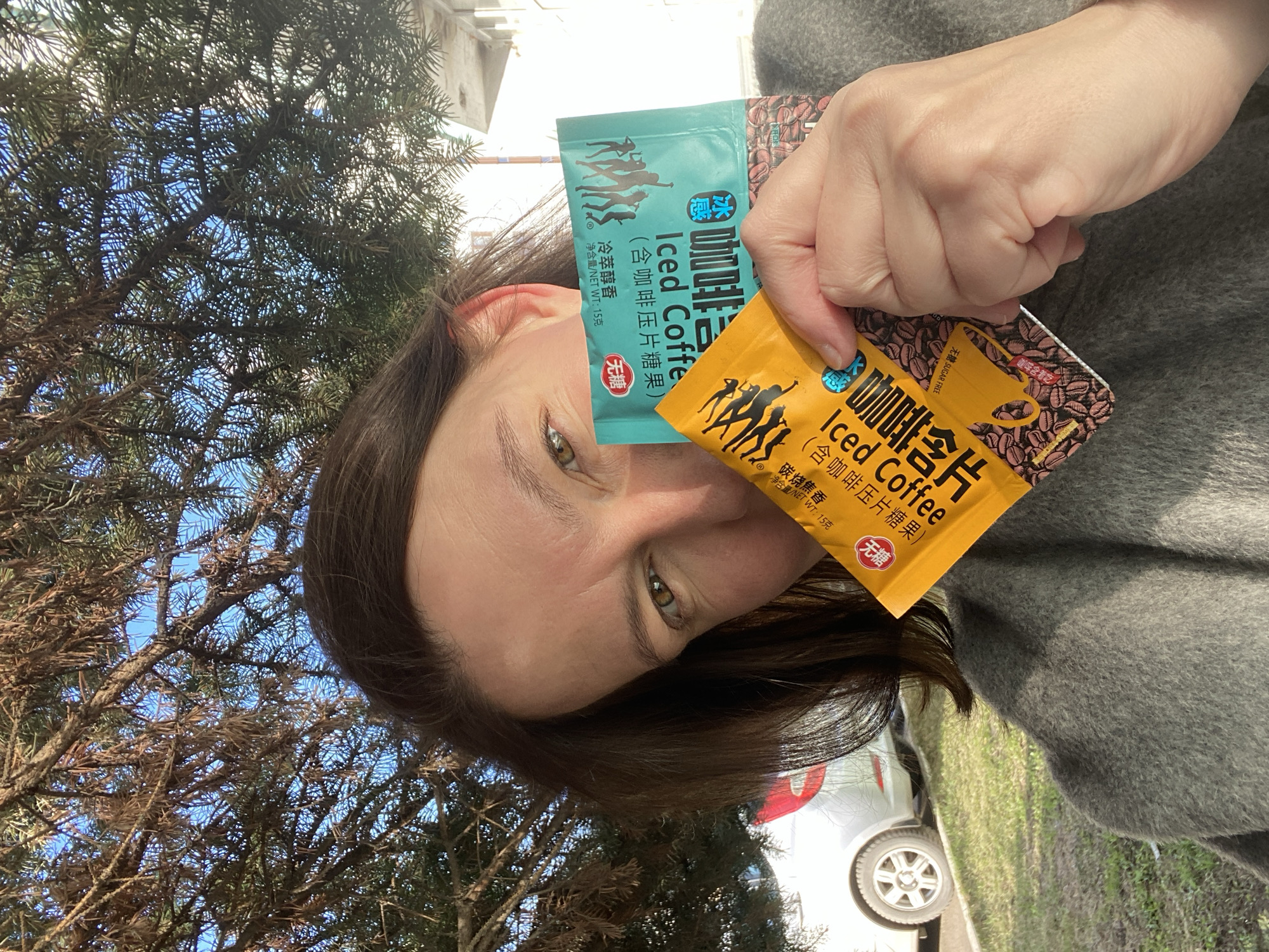 The image shows a woman holding two packets of "Iced Coffee" candy. The packets are colored teal and yellow. The woman has brown hair and green eyes. She is wearing a gray jacket. The background includes a tree with sparse branches and a glimpse of a silver car and buildings, suggesting an outdoor setting. The city of Chaikovsky, Russia, is not apparent in the image; this location data is not verifiable.