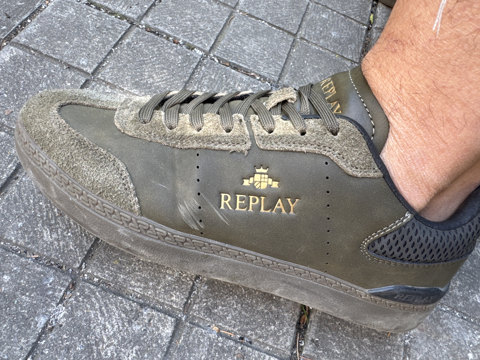 The image shows a close-up of a person's foot wearing a green "Replay" branded sneaker on a paved surface. The sneaker has a suede-like material on the toe and around the laces, with the "Replay" logo in gold lettering on the side. The sole of the shoe appears worn. The paved surface consists of grey square tiles. There is no specific information in the image to determine if the photo was taken in Plovdiv, Bulgaria.