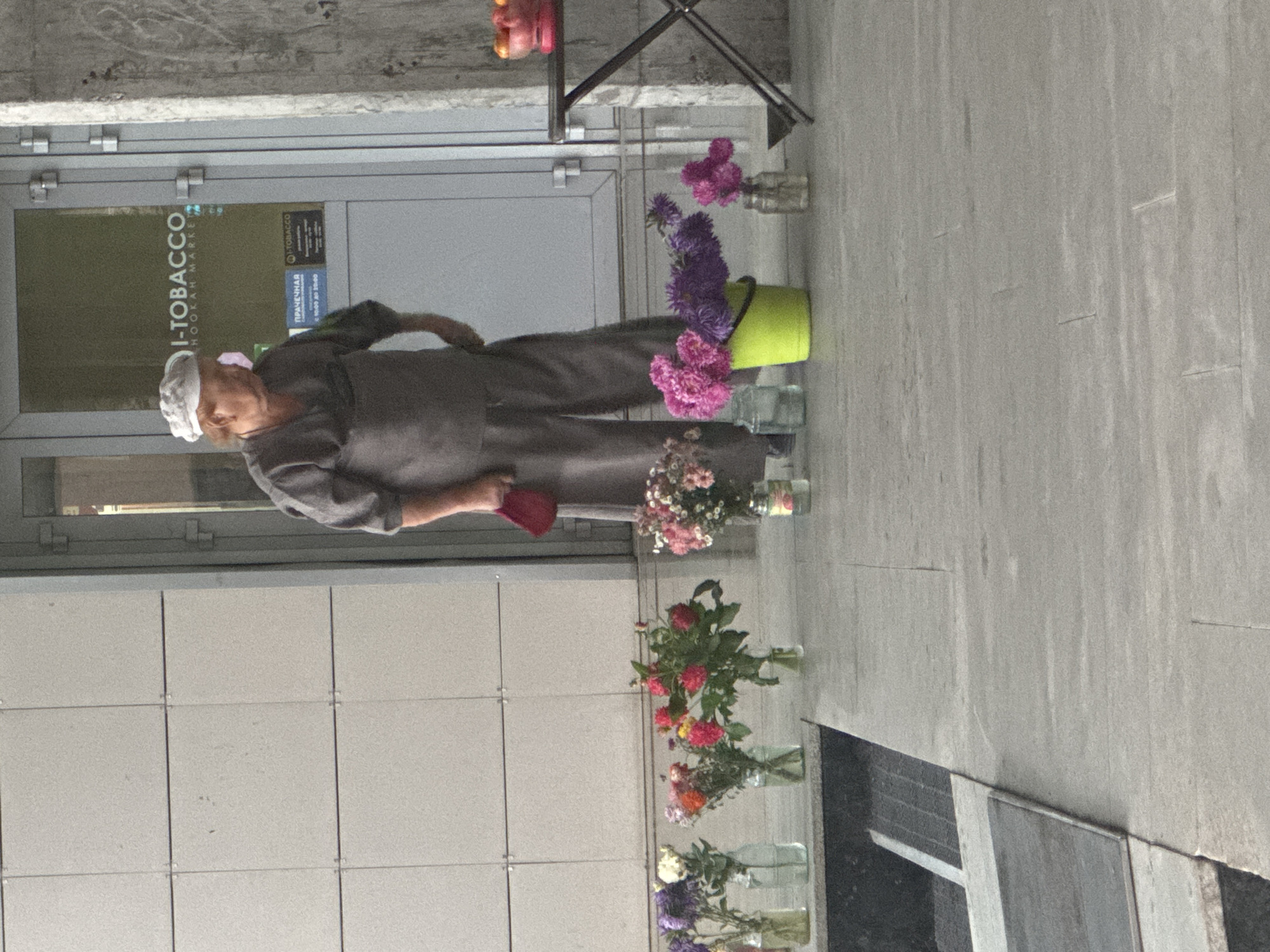 The image depicts an outdoor scene in Ufa, Russia, in front of a building with a glass door that has a sign for "I-TOBACCO HOOKAH MARKET". A person, appearing to be an elderly woman wearing a light-colored cap and a gray outfit, is standing to the right of the entrance. She is holding a red object in her right hand. In front of the building, a row of glass jars and a bright green bucket contain various bouquets of flowers. Some of the flowers are pink, purple, and red. To the far right, a small table with a dark metal frame holds a pink bowl filled with oranges. The ground is paved with large gray tiles. The overall environment suggests a commercial area, possibly a street or a public walkway.