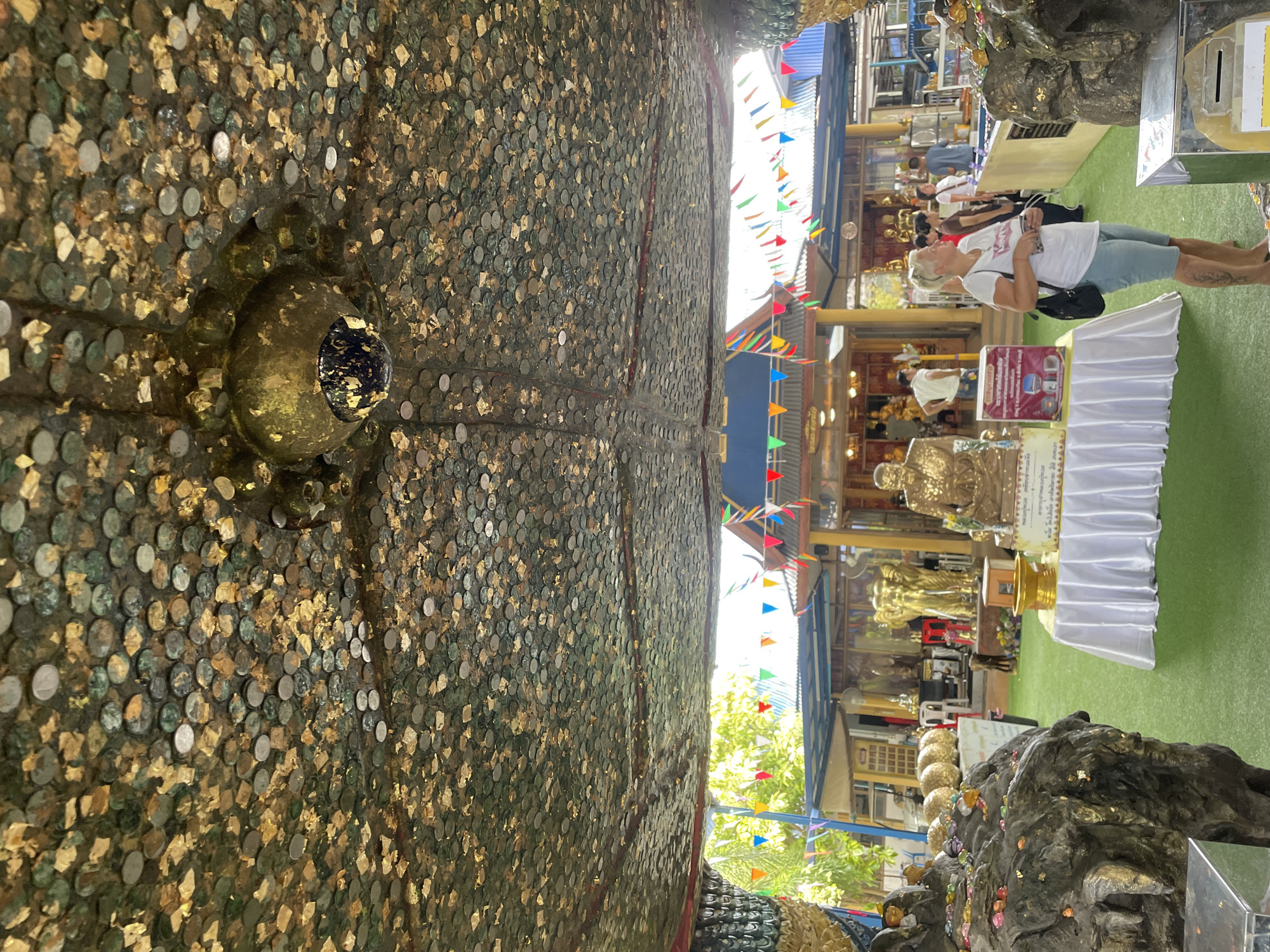 The image depicts an outdoor temple area, likely in Tambon Na Chom Thian, Thailand, characterized by a green artificial turf ground and structures adorned with Buddhist iconography. A prominent feature is the ceiling, which appears to be covered in a mosaic of coins and gold leaf, creating a shimmering effect.
In the foreground, to the left, a large, dark, rocky structure is visible, also partially covered in gold leaf and small colorful offerings. To the right, a white-clothed table is set with a golden bowl and what appears to be a ceremonial offering box or shrine. A golden statue of a seated monk is positioned behind the table.
In the mid-ground, several golden statues are displayed, including what appears to be two large elephant statues and more monk statues. A white-clad woman with blonde hair, wearing a white t-shirt and light blue shorts, is standing in the mid-right of the frame, looking towards the offerings. Several other individuals, appearing to be tourists or visitors, are scattered in the background, some observing the statues and others interacting with the temple environment. Colorful triangular flags are strung across the upper portion of the scene, adding a festive atmosphere. The background reveals more of the temple buildings, with various architectural elements and more decorative flags. The overall environment suggests a place of worship and cultural significance, likely a tourist attraction within Tambon Na Chom Thian.