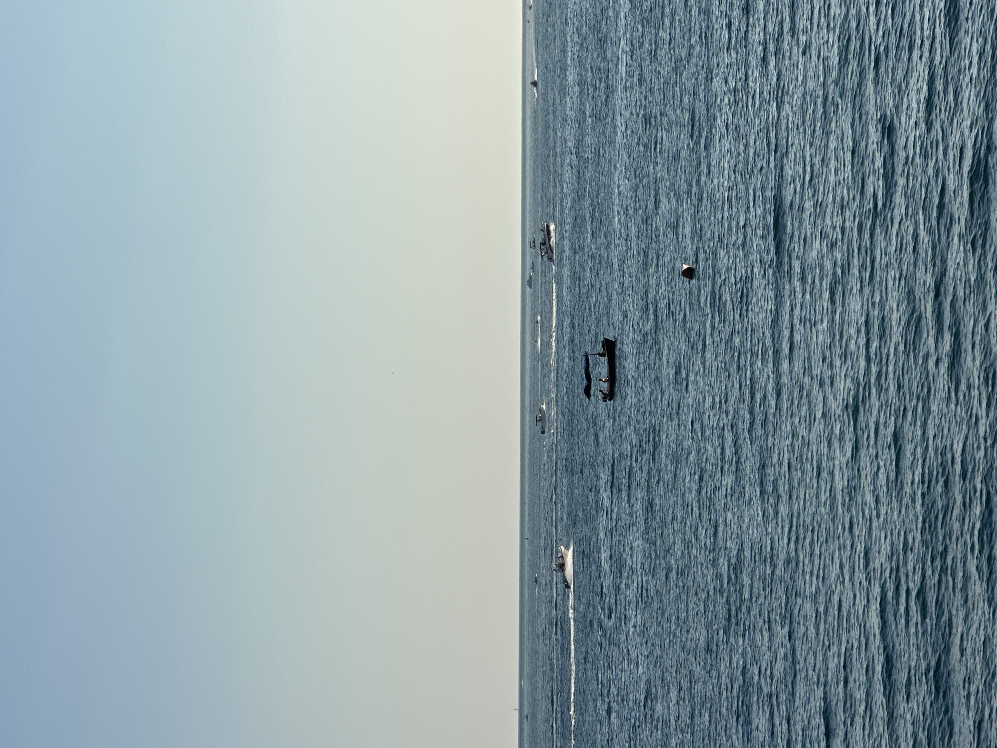 The image depicts a wide expanse of calm, deep blue sea under a pale, hazy sky. Several boats are scattered across the water, some appearing to be speedboats leaving white wakes, suggesting movement. In the foreground, a small, dark boat with a blue canopy is visible, carrying at least one person. Further out, other boats are seen, with one notable speed boat moving from left to right, leaving a prominent white trail. A red and white buoy is floating in the middle ground. The scene appears to be a sunny day with clear visibility. Given the context of Budva, Montenegro, this scene likely captures a view of the Adriatic Sea, possibly near the Budva Riviera, where boat activity is common.