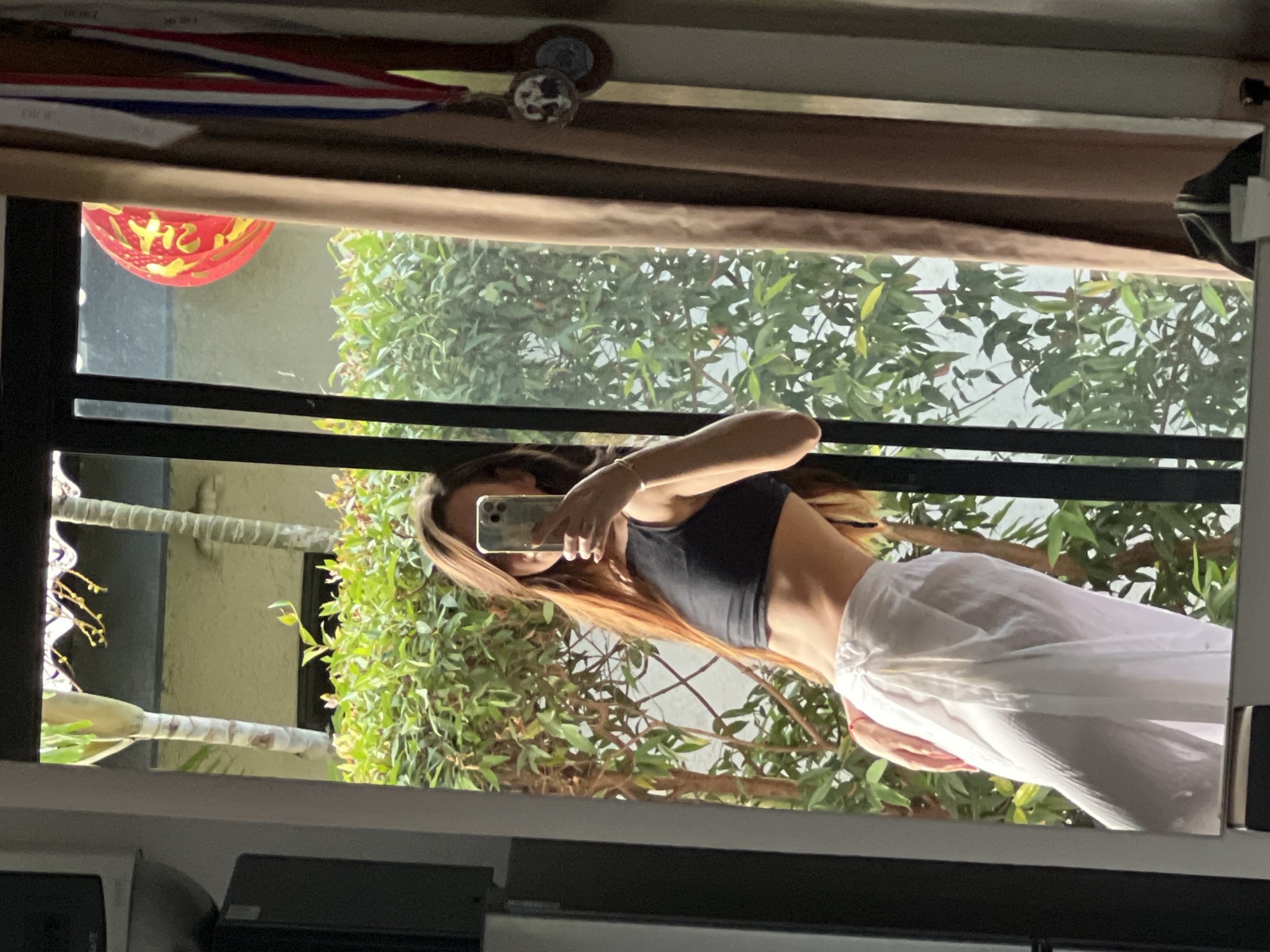 A mirror reflects a person in a black top and white pants holding a smartphone. The reflection includes greenery and a building exterior. A red lantern is visible near the top left of the mirror. The location is Unknown, Unknown.