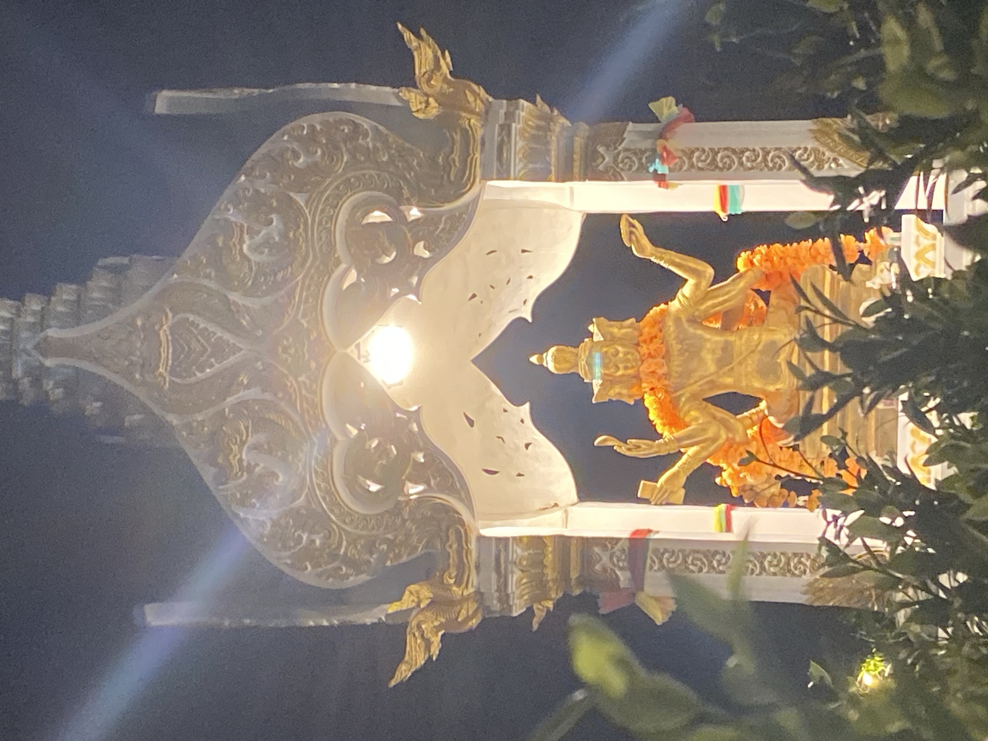 The image displays a golden, multi-headed (at least three visible) and multi-armed (four visible) statue, likely depicting Phra Phrom (Brahma), adorned with vibrant orange floral garlands. The statue is seated within an ornate, multi-tiered white and gold shrine, consistent with Thai architectural styles. A single, bare incandescent light bulb is suspended directly above the statue, casting a strong, warm glow that illuminates the figure and the interior of the shrine. This illumination creates distinct lens flares, including a prominent rainbow-colored streak in the upper left and a diagonal beam in the lower right. The shrine is supported by four pillars, decorated with golden sculptural elements resembling mythical birds or garudas, and wrapped with colorful fabric strips (red, yellow, green, blue). The scene is set at night, with a dark sky forming the background. Lush green foliage partially frames the lower and side edges of the scene, with some leaves appearing closer to the camera and slightly out of focus. The location is in บ้านบางรักษ์, Thailand.