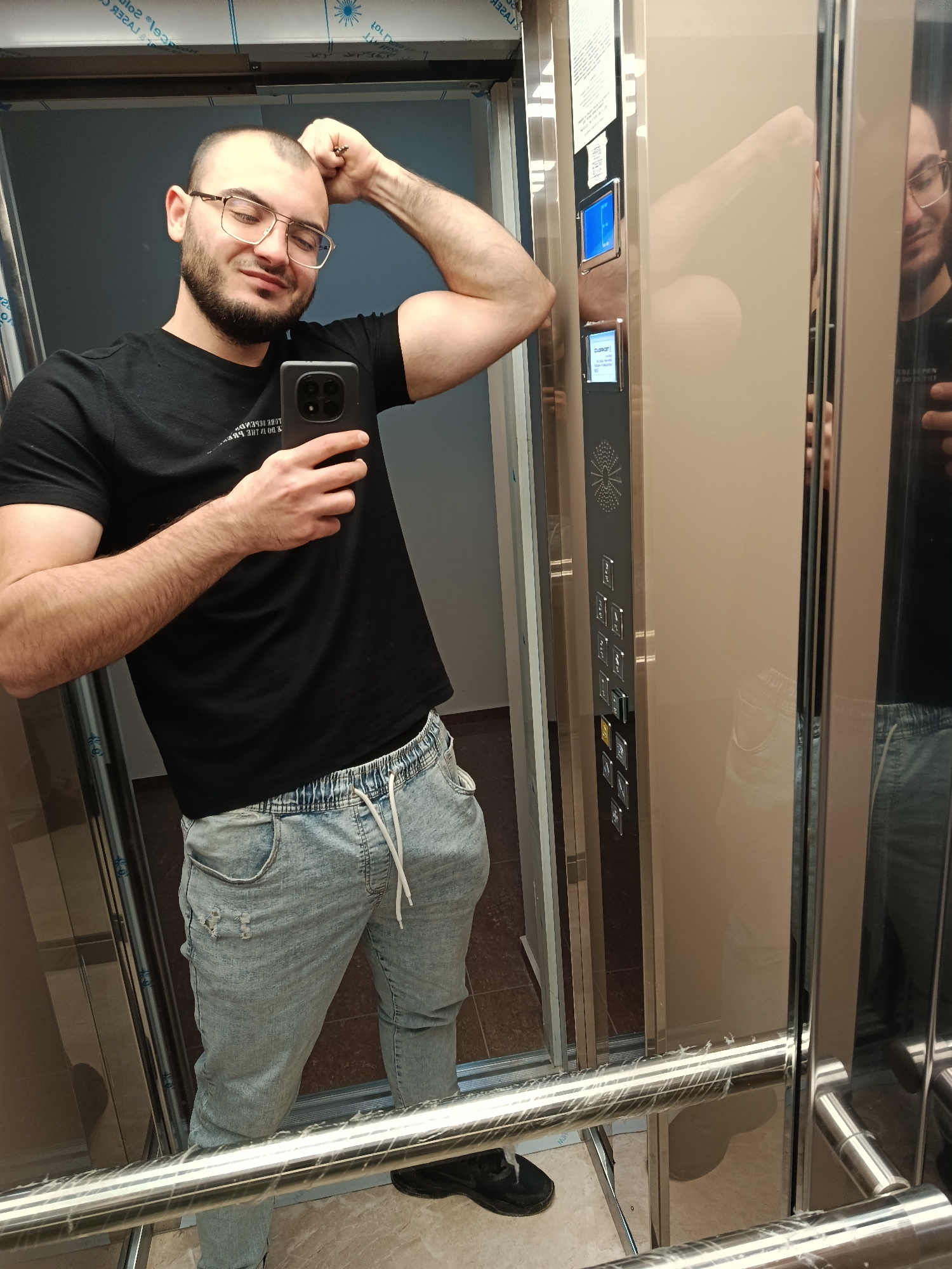 A man is taking a selfie in an elevator. He is wearing a black t-shirt, light blue ripped jeans, and black shoes. He is holding a smartphone in his left hand and has his right arm raised, flexing his bicep. He has short brown hair, a beard, and is wearing glasses. The elevator has a mirrored wall reflecting the man and the elevator's control panel. The elevator interior is beige with silver accents. The floor appears to be tiled in a light brown color. The location is possibly within a building in Plovdiv, Bulgaria.