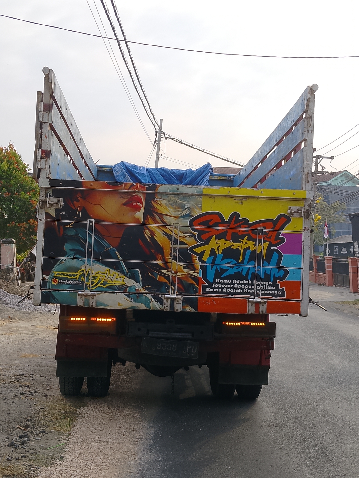In the city of Ngelo, Indonesia, a rear view of a brightly decorated flatbed truck is captured. The truck is adorned with a large graphic on its side featuring a woman's face, her lips painted red, and her hair blowing in the wind. Below the image of the woman, text in yellow and white is visible. The truck's tailgate is also decorated with colorful graffiti-style lettering in red, orange, yellow, green, and purple. Two lines of text in white are written below the graffiti, with the top line appearing to say "Kamu Adalah Dunianya" and the bottom line "Kamu Adalah Karyawannya". The truck has orange brake lights and a license plate with the characters "9358 JPD". The truck is driving on a paved road, with a dirt and gravel shoulder on the left side of the image. Trees and buildings are visible in the background. The sky is overcast.