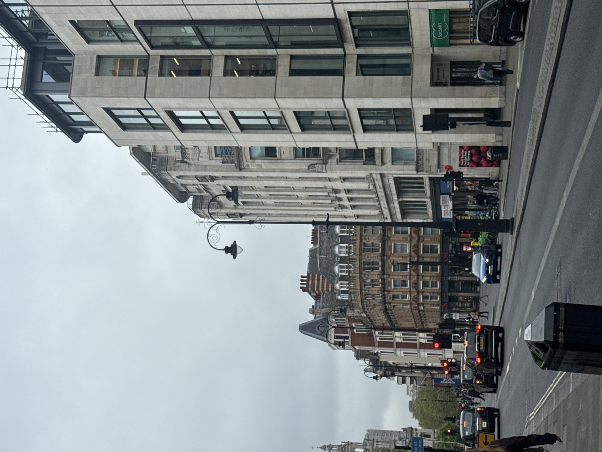 The image depicts a street scene in London, United Kingdom, under an overcast sky. The street is lined with multi-story buildings of varying architectural styles, ranging from modern to classical. A lamppost with decorative ironwork is visible in the middle ground. Traffic, including cars and black cabs, moves along the right side of the frame. Pedestrians are present on the sidewalks, and street furniture, such as benches and planters, is also visible. Signage and storefronts suggest a commercial area. The overall atmosphere is urban and somewhat muted due to the weather.