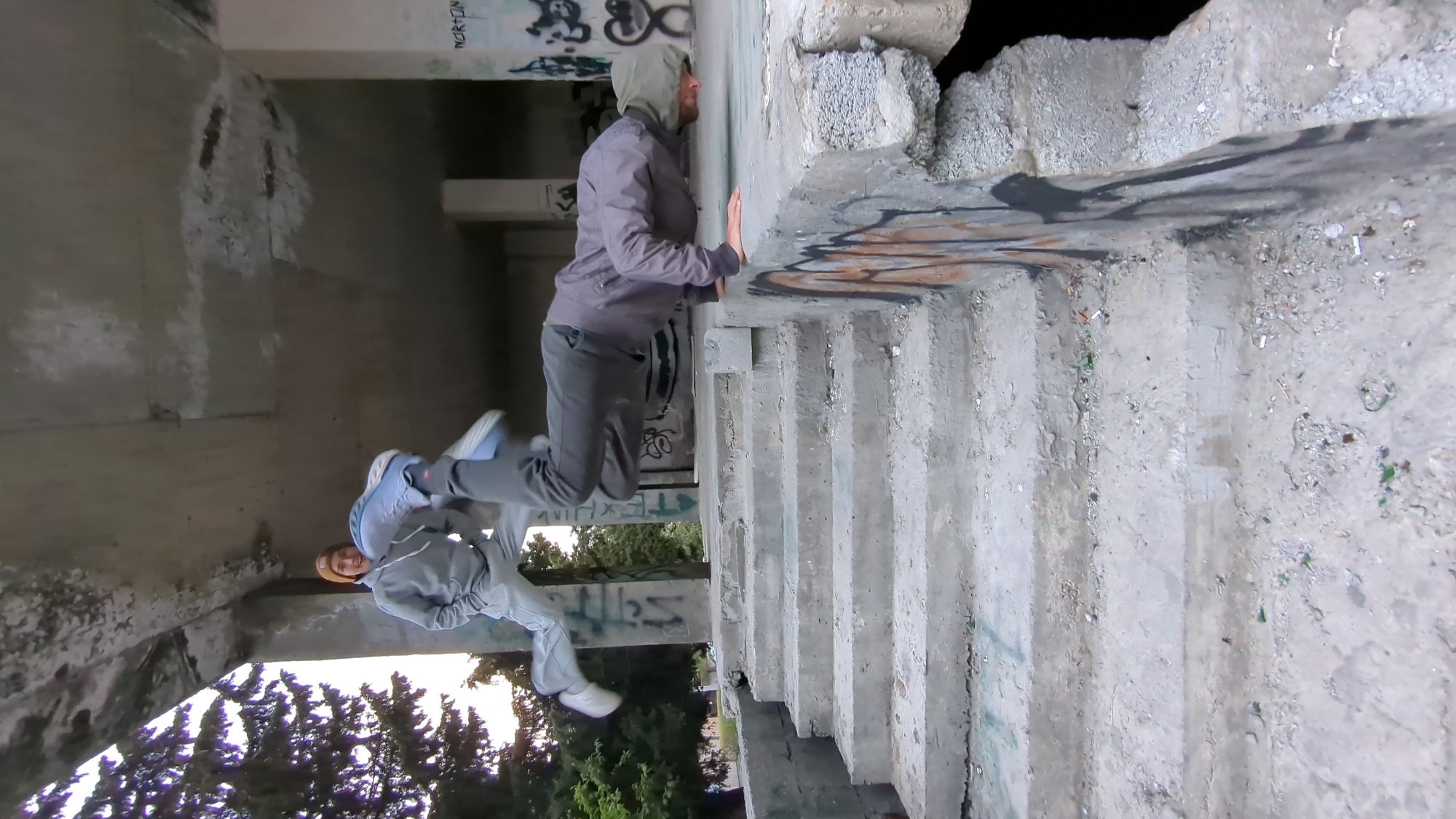 The image depicts an outdoor scene, likely in an abandoned or derelict structure. The location is reported to be in Gelendzhik, Russia. Two individuals are present; one appears to be jumping or leaping while the other seems to be balancing on a ledge or wall. The structures consist of concrete walls, pillars, and stairs, exhibiting signs of decay and graffiti. Some foliage, including trees, is visible in the background, suggesting the location is in an area with vegetation.