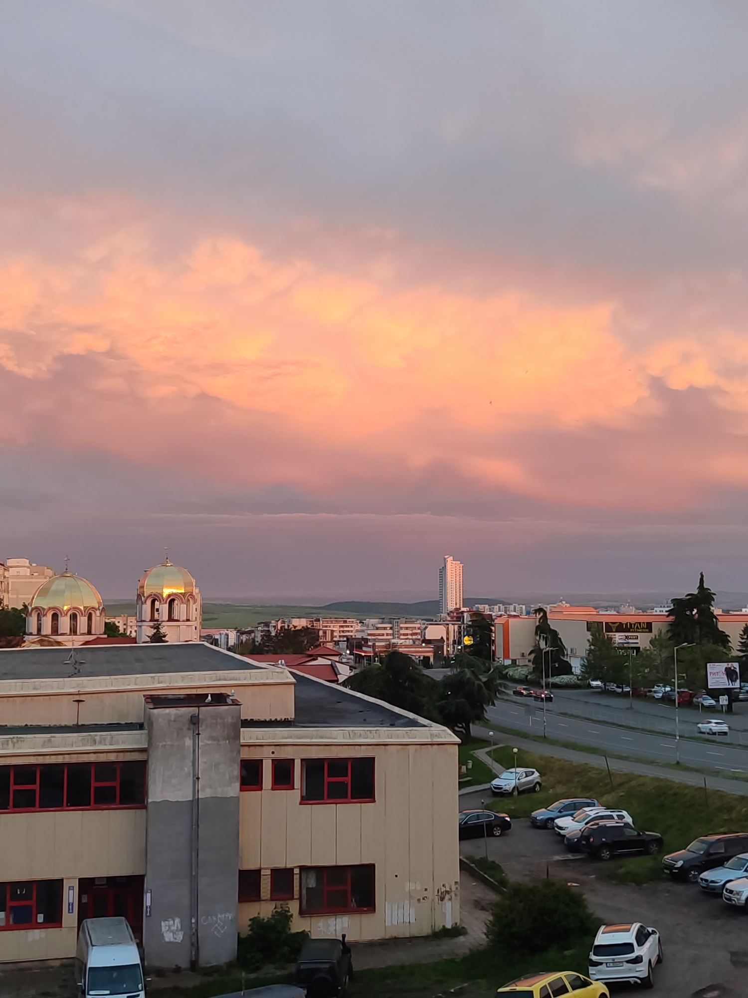 The image shows a cityscape under a sunset sky with pink and orange clouds. In the foreground is a beige building with a gray central section, red-framed windows, and some graffiti on the gray section. A white van is parked in front of the building. Beyond the building are streets with parked and moving cars. In the middle distance, golden dome roofs of a building are visible, alongside other buildings. A tall white skyscraper is visible in the background. The location is Burgas, Bulgaria.