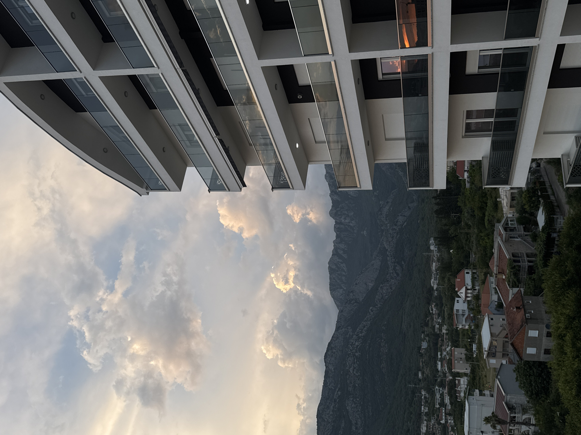 This media file depicts a view of a modern building with balconies overlooking a mountainous landscape and a residential area. The building is white with dark accents and glass railings on the balconies. The sky is filled with dramatic clouds, suggesting either sunrise or sunset, with hints of orange and pink reflecting on the building's windows.
The scene is set in Šušanj, Montenegro, as indicated by the presence of houses with terracotta roofs nestled on the hillside, characteristic of Mediterranean architecture. The dense green foliage and the imposing, rocky mountains further confirm the coastal, mountainous terrain. The overall mood is serene and picturesque, capturing a moment of natural beauty juxtaposed with modern architecture. No people are visible, and there is no visible text in the image. The time of day appears to be either early morning or late evening, indicated by the warm tones in the sky.