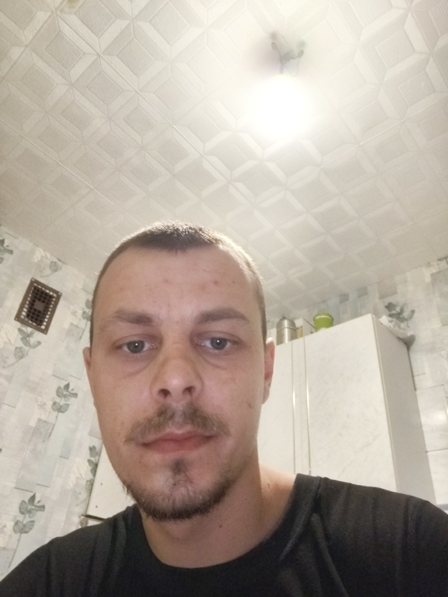 A close-up, low-angle selfie shows a man with short brown hair and a short beard and mustache looking directly at the camera. He is wearing a dark t-shirt. Behind him, a white tiled ceiling with a square pattern is visible. To his left, a rectangular metal vent is mounted on a wall with a light-colored floral wallpaper. To his right, a white cabinet is partially visible, with two containers on top of it. The background suggests an indoor residential setting, likely a kitchen or bathroom, consistent with a location in Коханаўскі сельскі Савет, Belarus.
