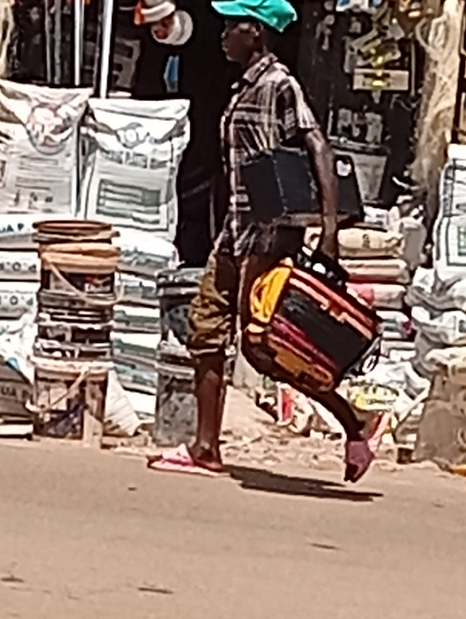 The image shows a person walking along a dusty street in Maiburiji, Nigeria, under bright daylight. The individual, appearing to be a man, is captured mid-stride, moving from left to right across the frame. He wears a short-sleeved plaid shirt, knee-length shorts, a bright green cap, and pink flip-flops. A dark, rectangular bag is slung over his left shoulder, and he carries a larger, colorful rectangular object with yellow, red, and black stripes in his right hand, possibly a portable music player or another type of bag.
The scene suggests a bustling commercial area, likely a market or a street lined with shops. On the left side of the frame, there are tall stacks of large white sacks, some partially visible with text such as "P.O.", "QUA P.", and "10KG". Alongside these sacks are stacks of plastic containers, including some yellow or orange buckets. The background on the right also features numerous bags and indistinct items, reinforcing the impression of a busy marketplace. Strong shadows are cast on the light brown ground, indicating a sunny day. The overall activity depicted is a person moving through a lively street environment.