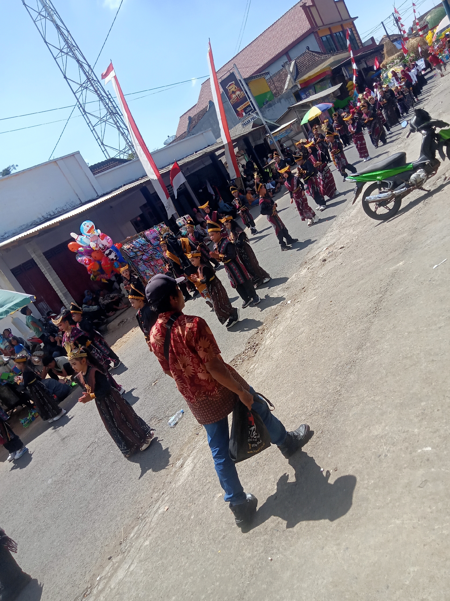 A parade is taking place on a street in Pule, Indonesia. A group of children in traditional attire are marching, followed by adults. A green motorcycle is parked on the side of the road. The sky is clear and blue, and buildings line the street. Flags and balloons are visible, adding to the festive atmosphere. The people are dressed in colorful clothing, and the children are wearing elaborate headdresses. The parade appears to be a cultural event, possibly a celebration of independence or a local festival. The overall scene is lively and engaging, with a sense of community and tradition.