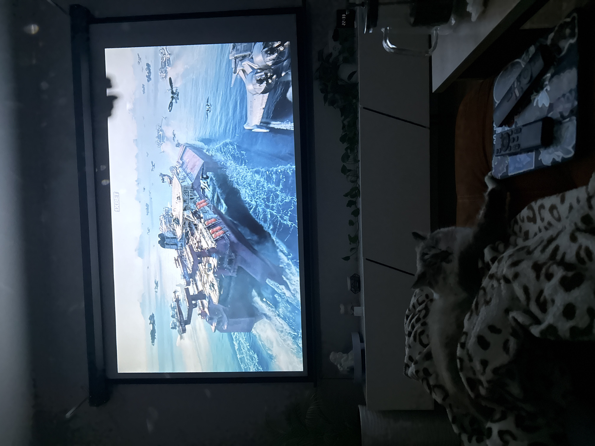 A rectangular projector screen displays an aerial battle scene on the left side of the image. The scene appears to be from a science fiction setting, featuring aircraft and naval vessels. To the right of the screen, partially visible, are some objects. A cat rests on a patterned blanket. On the top of a white shelf, remotes are placed on a flowered cover. A glass and some decorative objects are also present on the shelf. The time 22:33 is visible on a small, dark device on the wall. The environment is dimly lit. There is no evidence to suggest that this scene is located in Rostov-on-Don, Russia.