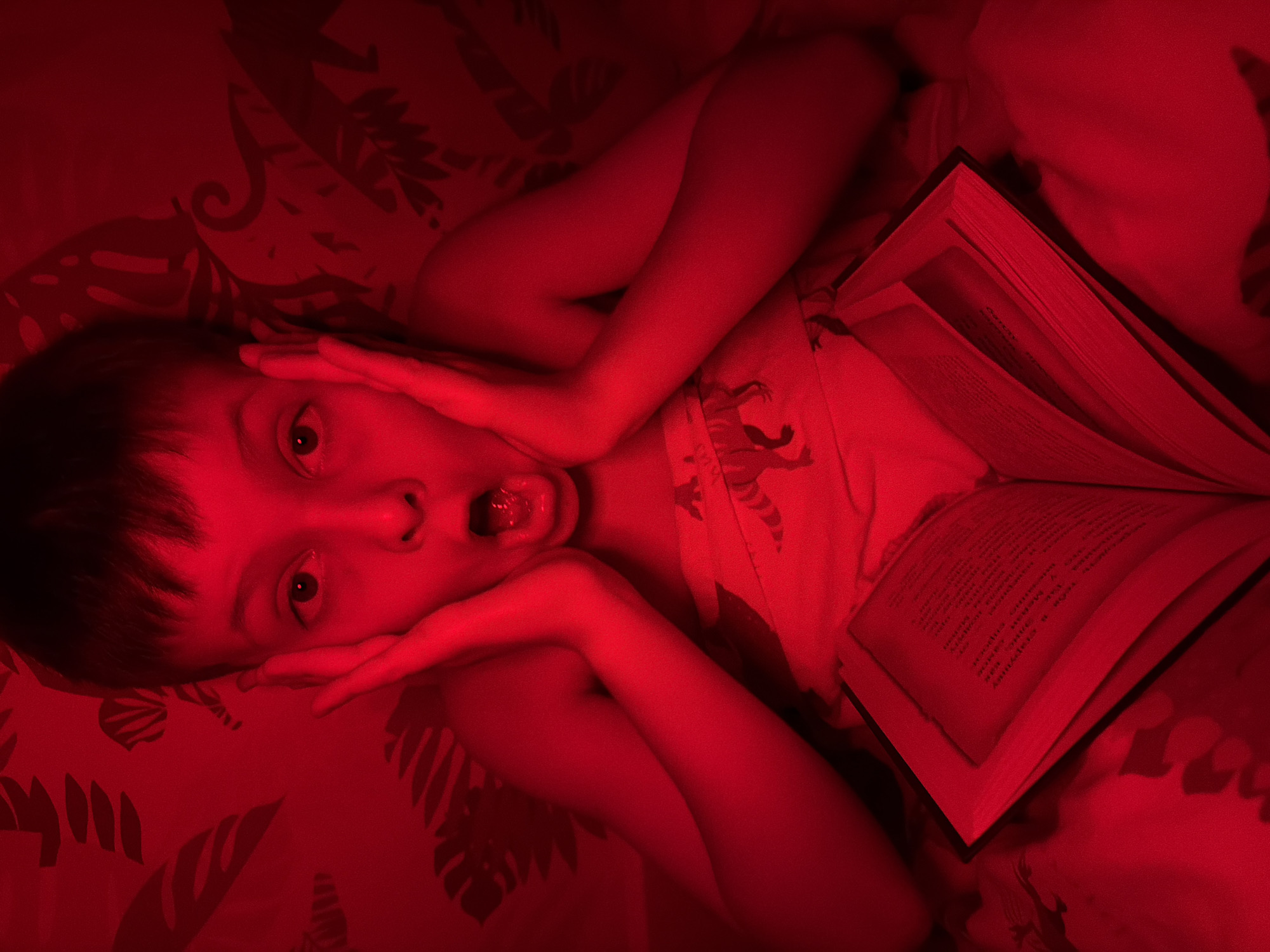 A child with short, dark hair lies on a bed under red lighting, positioned with their head tilted towards the upper left of the frame. The child's eyes are wide and their mouth is open, tongue visible, with both hands framing their face. A book is open beside the child, resting on the bed. The bedsheets feature a dark leafy pattern. The lighting is predominantly red, affecting the overall color balance of the image. There is no indication of the city of Votkinsk, Russia.