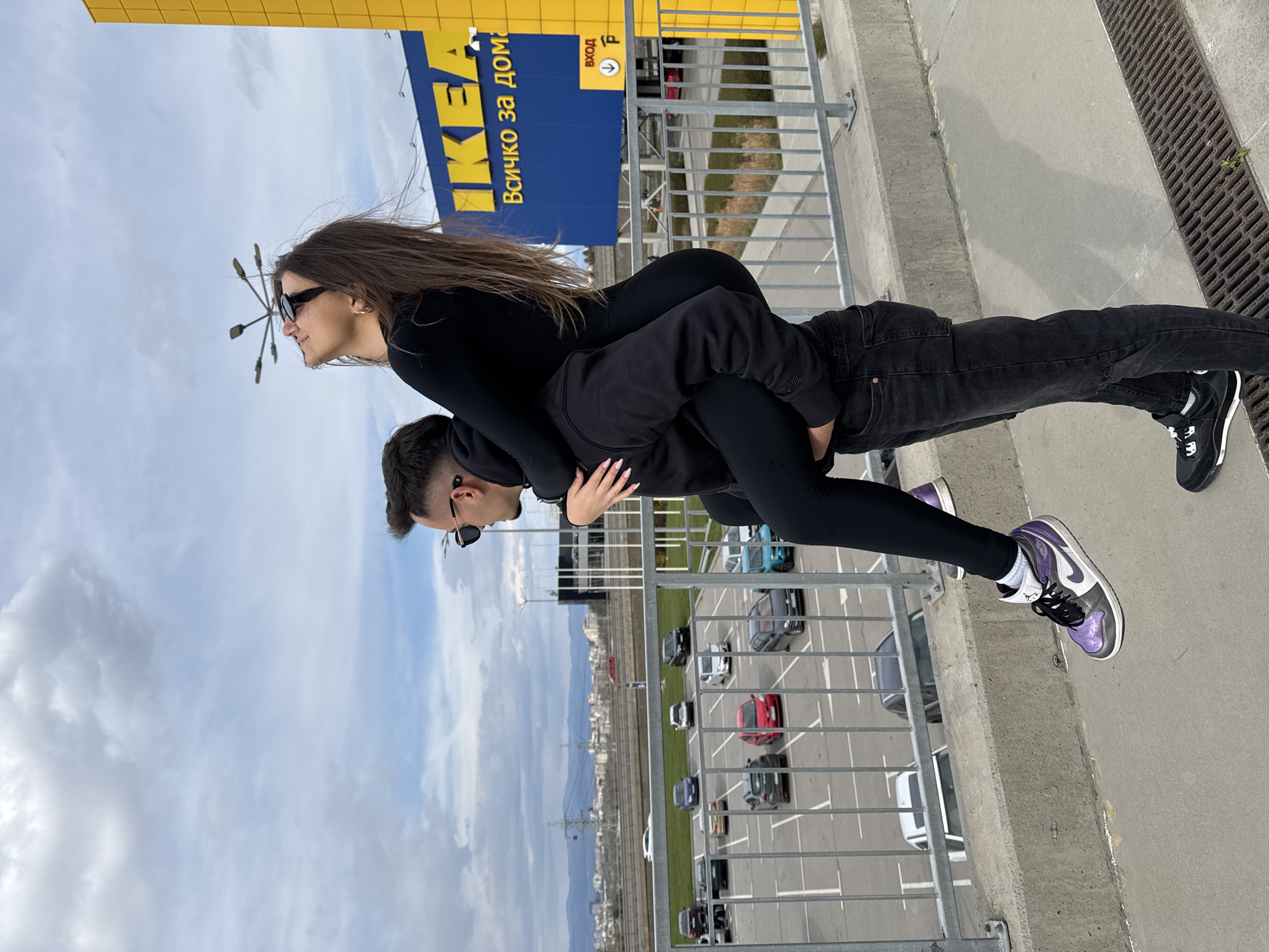 A man and a woman are posing for a photo on the roof of the IKEA store in Sofia, Bulgaria. The woman is riding piggyback on the man, with her arms around his neck. The man is wearing black jeans, a black hoodie, and black-and-white sneakers. The woman is wearing a black bodysuit and purple and white sneakers. Both of them are wearing sunglasses. In the background, there is a parking lot filled with cars and a cityscape with mountains in the distance. The sky is cloudy. The IKEA building is visible in the upper part of the frame.