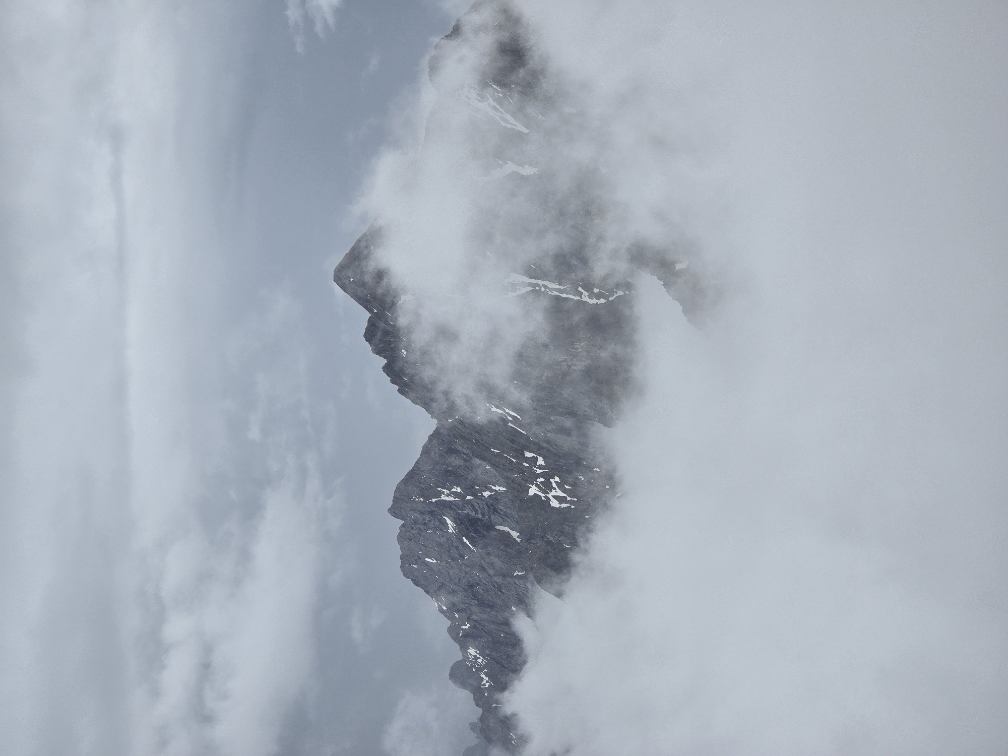 The image depicts a dramatic mountain landscape partially obscured by dense fog and clouds. The prominent subjects are rugged, rocky mountain peaks, some of which are dusted with patches of snow. The terrain is steep and appears to be made of dark grey rock. The fog creates a sense of mystery and height, with the lower portions of the mountains completely enveloped. The sky above is overcast and grey, with layers of clouds contributing to the muted atmosphere.
This scene is consistent with the natural environment surrounding Garmisch-Partenkirchen, Germany, a Bavarian town situated in the Alps. The Zugspitze, Germany's highest mountain, and other peaks in the Wetterstein range are known for their dramatic topography and susceptibility to fog and changing weather conditions, making this a plausible representation of the region's mountainous terrain. No people, structures, or human activities are visible in the image.