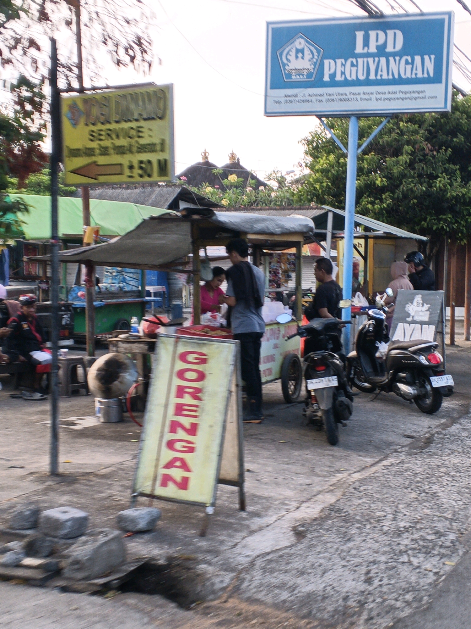 The image depicts a street scene in Denpasar, Indonesia, likely near a food stall area. In the foreground, a yellow sign with red text reads "GORENGAN," indicating fried food. To the left, a man in a black jacket with red accents sits on a stool. Behind him, a stall with a gray awning sells food items. Further back, a blue and green food cart is visible. On the right, a woman in a pink top is interacting with someone behind a stall. Two scooters are parked on the street.
Above the food stalls, a blue sign with white text reads "LPD PEGUYANGAN," which likely refers to a local credit union or financial institution. Beneath this sign, smaller text provides an address: "Alamat: Jl. Ahmad Yani Utara, Pasar Anyar Desa Adat Peguyangan." This confirms the location as the Peguyangan Village area, North Denpasar. A second yellow sign with black text, partially obscured by a pole, reads "YOGI DAMANS SERVICE," with an arrow pointing left and "± 50 M."
The environment appears to be an outdoor, possibly humid, setting with concrete ground and some foliage in the background. The overall impression is of a bustling local marketplace.