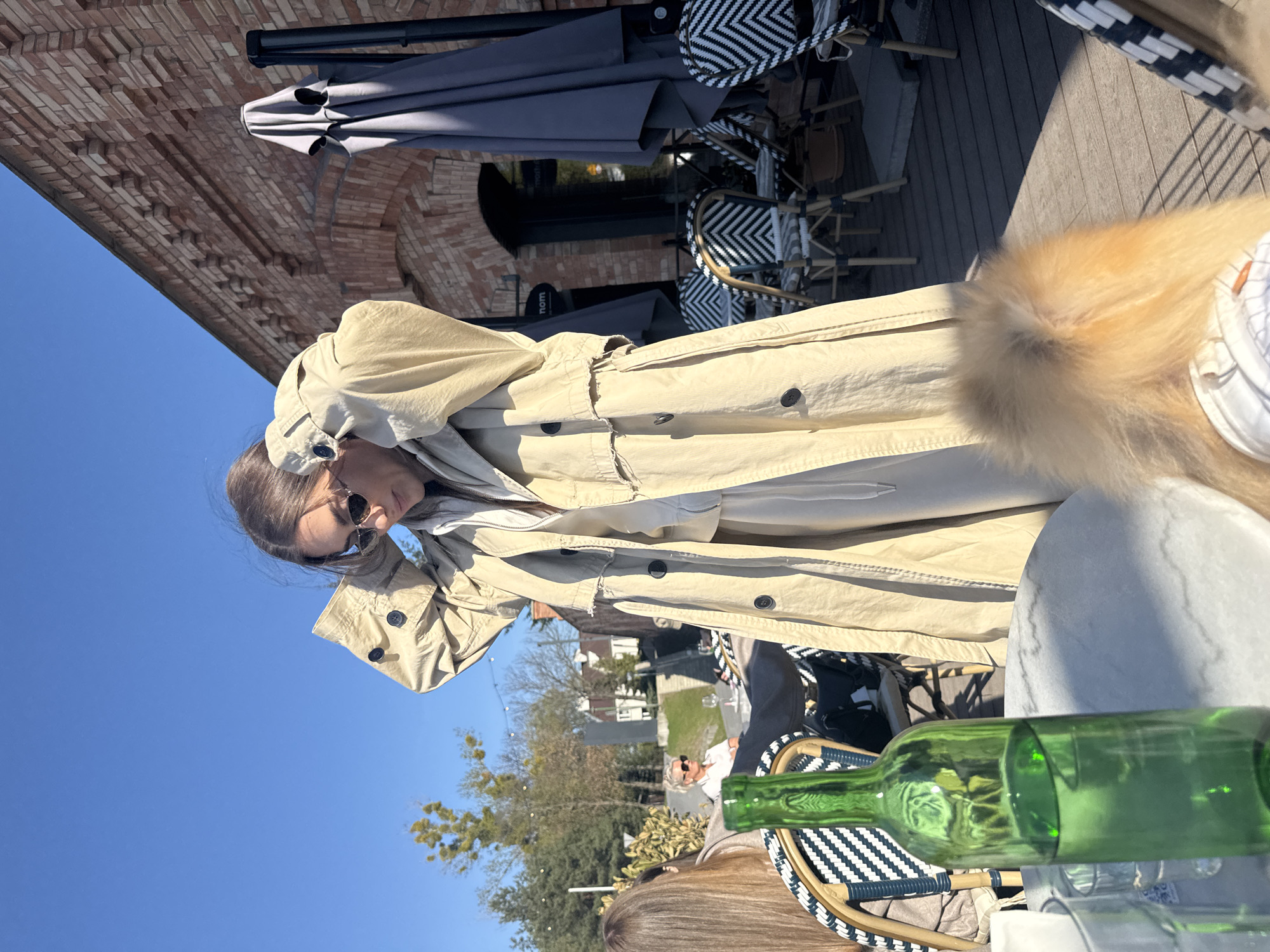 The image shows a woman wearing a tan trench coat and sunglasses sitting at an outdoor table at a restaurant. A partially visible green wine bottle sits on the table along with drinking glasses. Other people are seen at nearby tables. The outdoor restaurant features gray umbrellas and black and white patterned chairs. The restaurant is located in Warsaw, Poland, as indicated by signage on the restaurant. A brick building is also visible. The sky is blue with some trees in the background.
