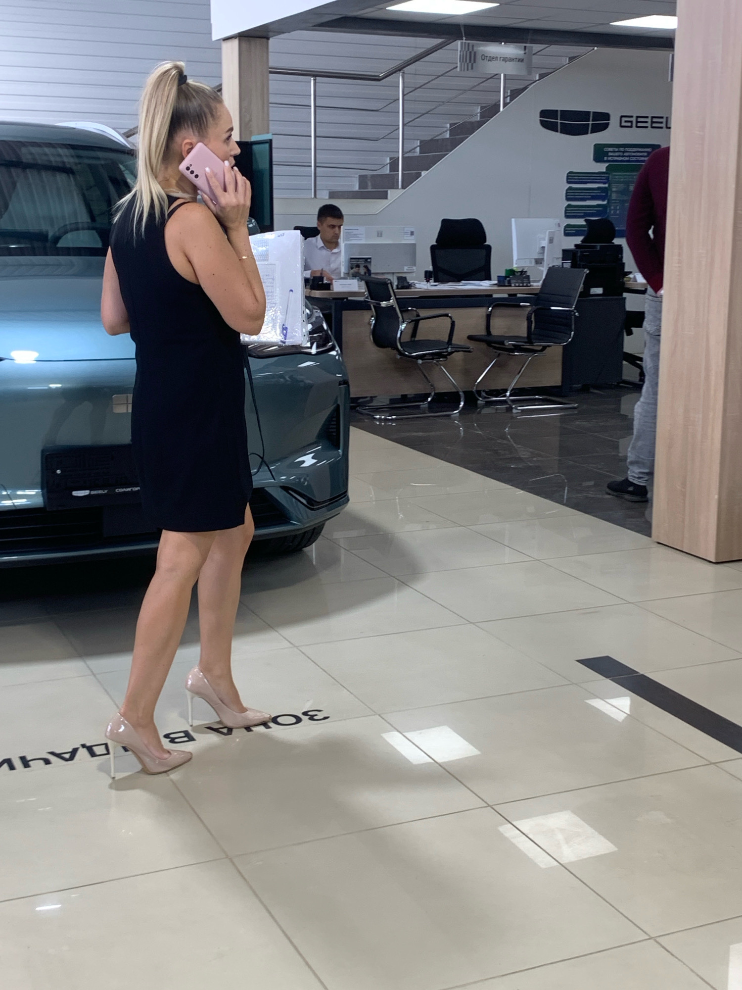 In a brightly lit showroom, a blonde woman with her hair in a high ponytail is on the phone, holding a pink smartphone to her ear. She is wearing a sleeveless black dress and high-heeled shoes. The woman is standing next to a dark green car. In the background, a man in a white shirt is seated at a desk, and another person in a maroon shirt and grey pants is visible. The showroom appears to be a car dealership, with desks, chairs, and signage, including a sign that reads "GEEL". The floor is tiled with a beige and white pattern. The overall environment suggests a commercial setting, possibly a car dealership located in or around Чыжэвіцкі сельскі Савет, Belarus, given the Russian language on the signs.