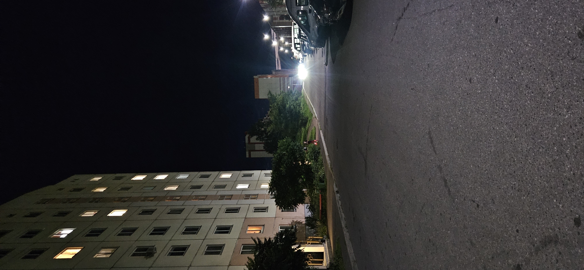The image shows a residential building in Vitsebsk, Belarus, at night. The building is tall and multi-story, with many windows illuminated from within. In front of the building is a paved road and a parking lot where several cars are parked. Trees and greenery line the side of the road. In the distance, other buildings and streetlights are visible. The sky is dark, indicating nighttime. A person is visible on the sidewalk in the middle ground, wearing dark clothing.