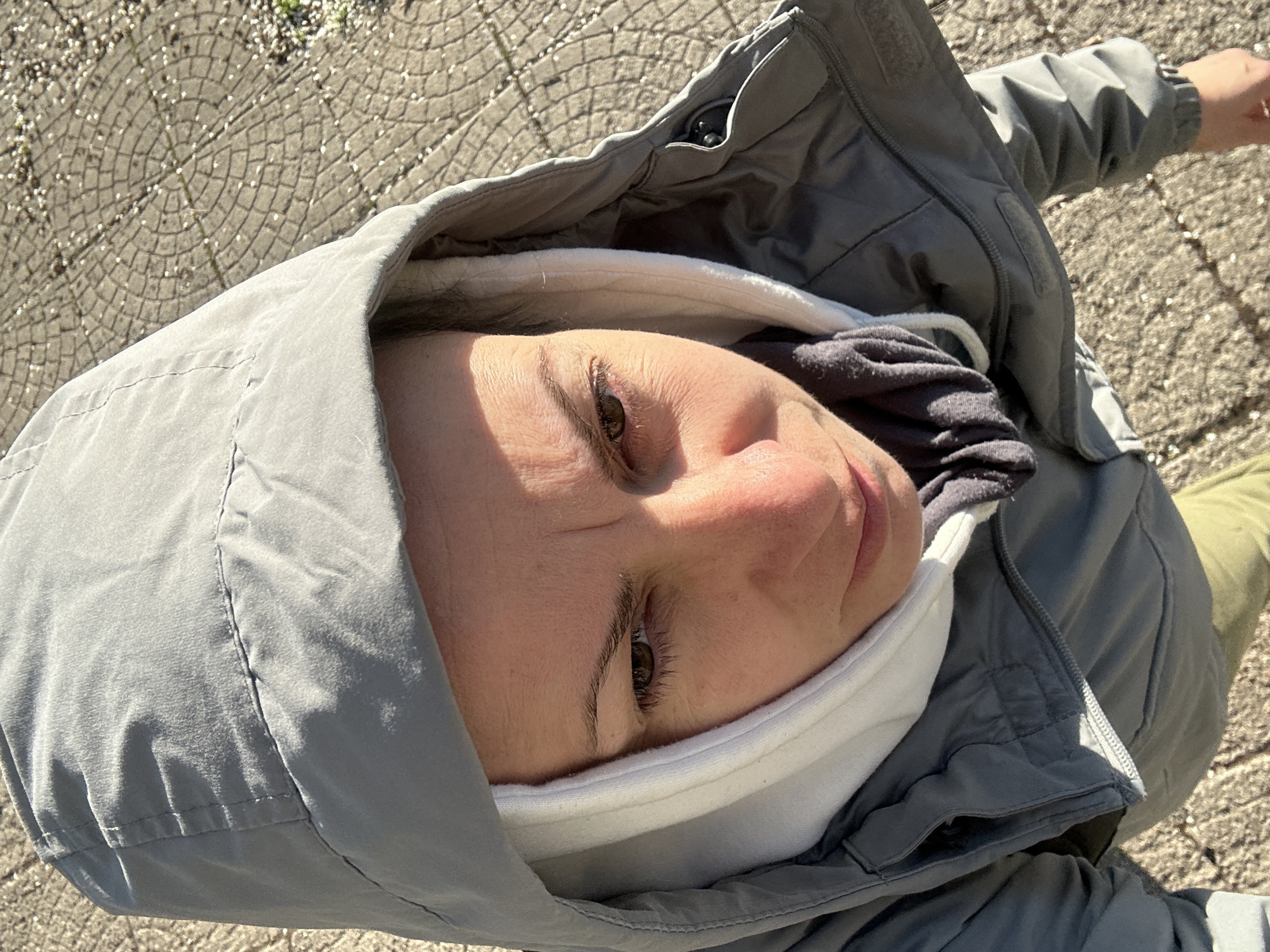 A woman is seen from a high angle, possibly taken by herself. She is wearing a grey hooded jacket with a white fleece neck warmer and a grey top visible underneath. The woman's face is in focus, showing her skin tone and brown eyes. She is looking directly at the camera. The background includes a patterned stone sidewalk. The exact location in the city of Sofia, Bulgaria cannot be determined.