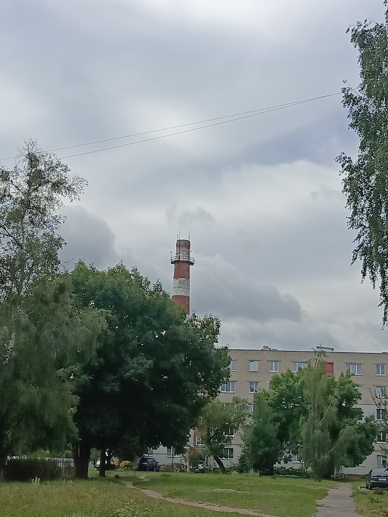 This image depicts a daytime scene in Ivatsevichy, Belarus. In the background, a tall, red and white striped industrial chimney rises against a cloudy sky. In front of the chimney, a multi-story apartment building, constructed from light-colored concrete blocks, features numerous windows and a single red-colored window or door on its upper level. The foreground is dominated by lush green trees and grass, with a paved walkway visible leading towards the apartment building. A few parked cars are situated in the vicinity of the building, and a dark-colored fence runs along the left side of the scene. The overall atmosphere is overcast and urban.