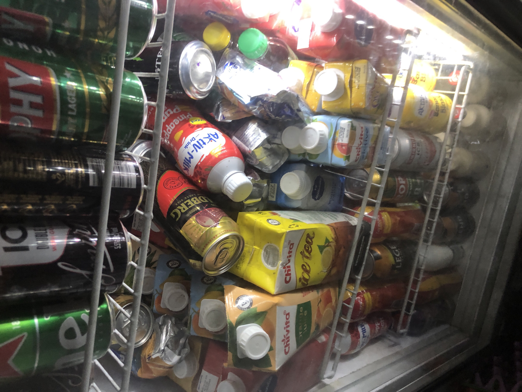 A refrigerated display case is packed with various beverages, including canned drinks and juice cartons. The refrigeration unit is well-lit, casting a bright glow on the products. The scene appears to be in a commercial establishment, likely a store or a bar, located in Ado, Nigeria.