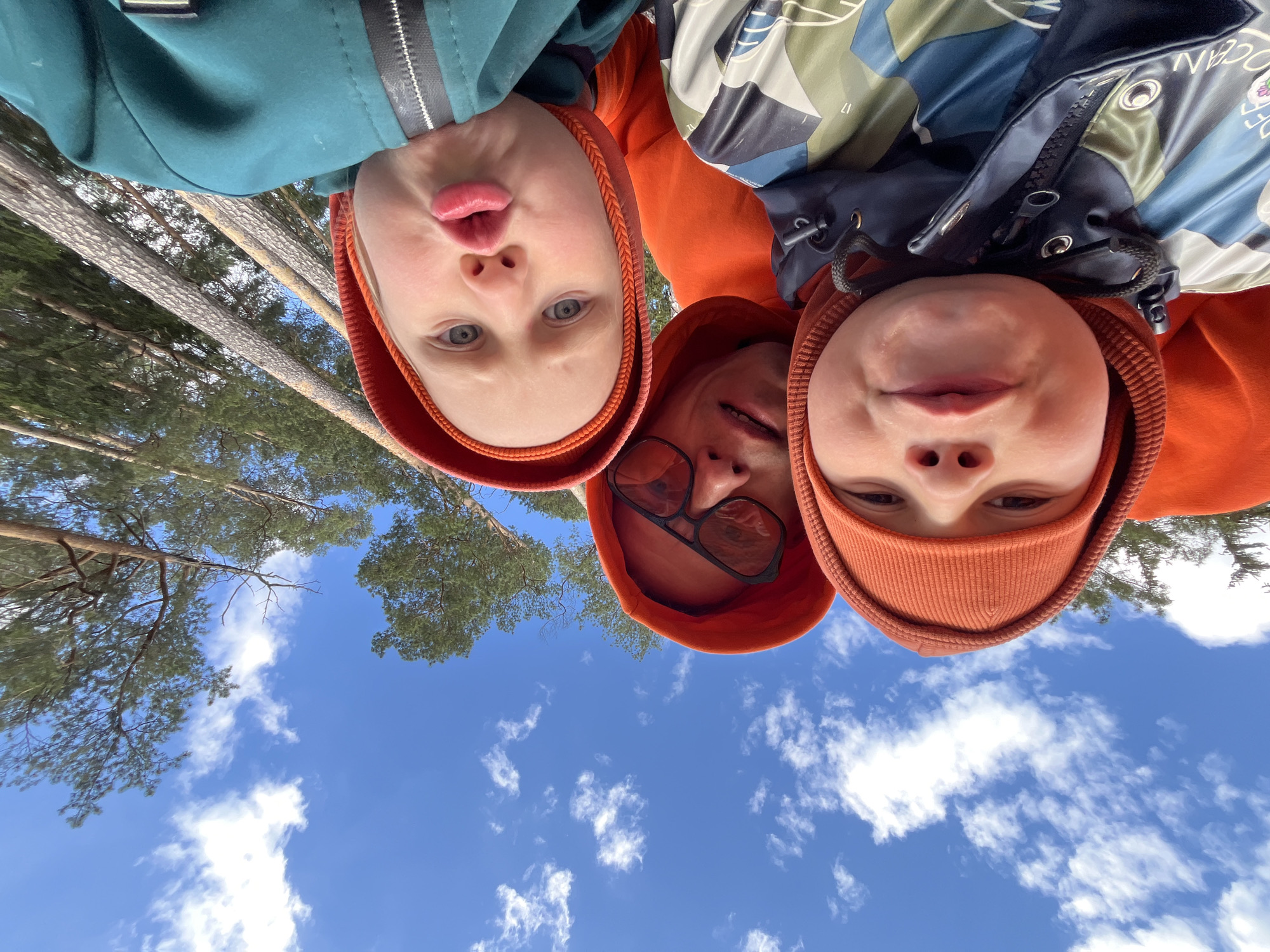 Three people are looking down at the camera, positioned in the center. They appear to be in an outdoor location, judging by the blue sky with scattered clouds visible in the background. Above and slightly to the sides of the subjects are pine trees. The person on the left wears a teal jacket with a red-orange hooded undershirt. The person in the center wears an orange hooded sweatshirt and eyeglasses. The person on the right wears an orange hooded sweatshirt and orange hat. The city of Chaikovsky, Russia cannot be determined from the information provided.