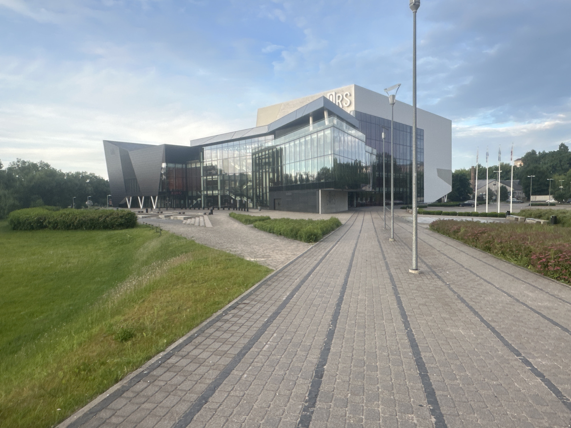 This image captures the modern GORS Concert Hall in Rezekne, Latvia, on a bright, cloudy day. The building's architecture is striking, featuring a combination of dark, angular geometric shapes and a large, transparent glass facade that reflects the sky and surrounding greenery. The name "GORS" is prominently displayed in large, white letters on the upper portion of the building.
In the foreground, a paved walkway made of grey interlocking stones with dark grey dividing lines leads towards the building. To the left of the walkway, a lush green grassy slope slopes downwards, dotted with some wild grasses and small flowering plants. Further to the left, manicured green bushes form a low hedge.
The walkway is lined with several tall, slender lamp posts. In the middle ground, beyond the glass facade, more greenery and trees are visible. To the right of the building, a row of flagpoles stands, and beyond them, other buildings and trees can be seen on a slight incline, suggesting the building is situated within a developed area. The sky is a mix of blue and scattered grey clouds, indicating fair weather. No people are visible in the scene.