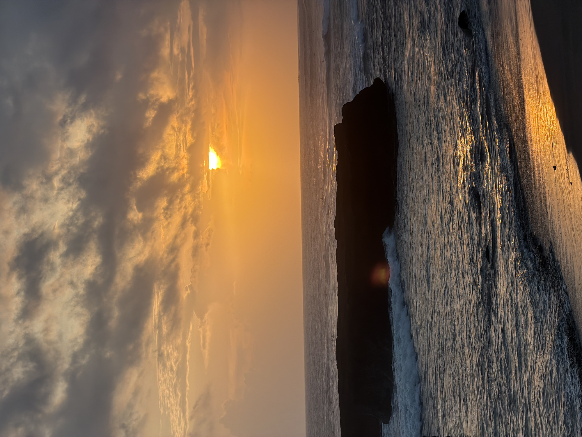 The image depicts a sunset scene in North Kuta, Indonesia. The sun is partially obscured by clouds, casting a warm orange glow across the sky. To the right, a rocky outcrop extends into the ocean, with waves crashing against its base. The beach stretches along the coastline, with wet sand reflecting the sunlight. There are no people visible in the image.