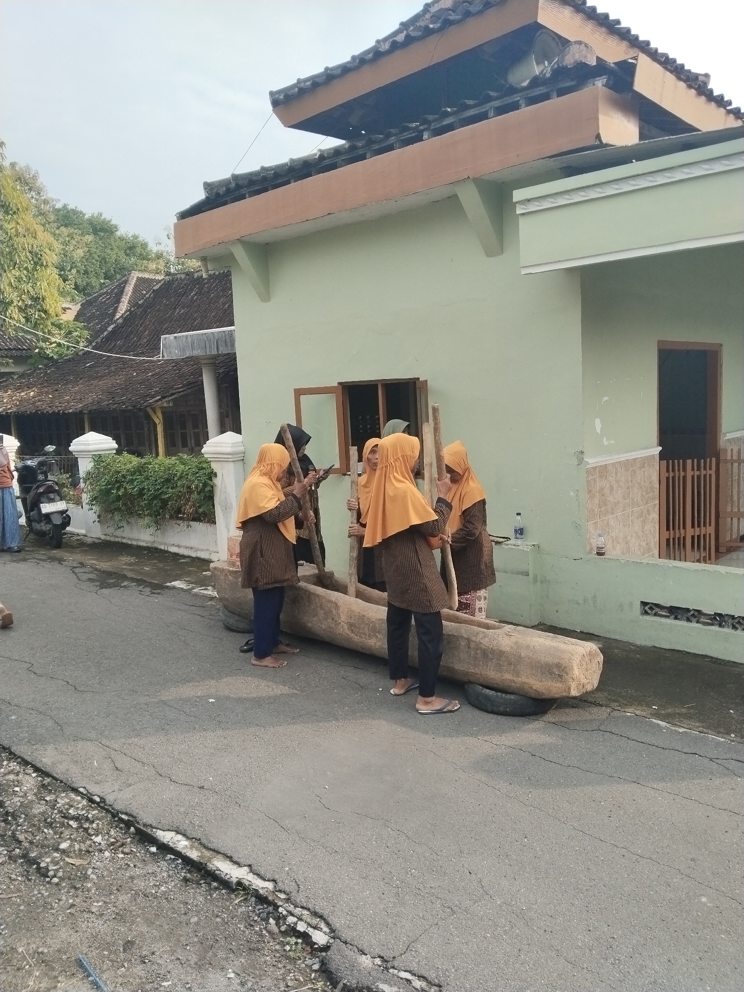 The image depicts a scene on a street in Gebang, Indonesia. A group of four women, all wearing yellow hijabs and striped shirts, are standing around a large, hollowed-out log placed horizontally on what appears to be a tire. Each woman is holding a wooden pestle, suggesting they are engaged in a grinding or pounding activity. The location is along the side of a light green building with a brown-framed window. In the background, there's another building with a dark-tiled roof, partially obscured by greenery and a parked motorcycle. The street itself is paved but shows signs of wear and tear. The overall impression is of a traditional or communal activity taking place in a residential area.