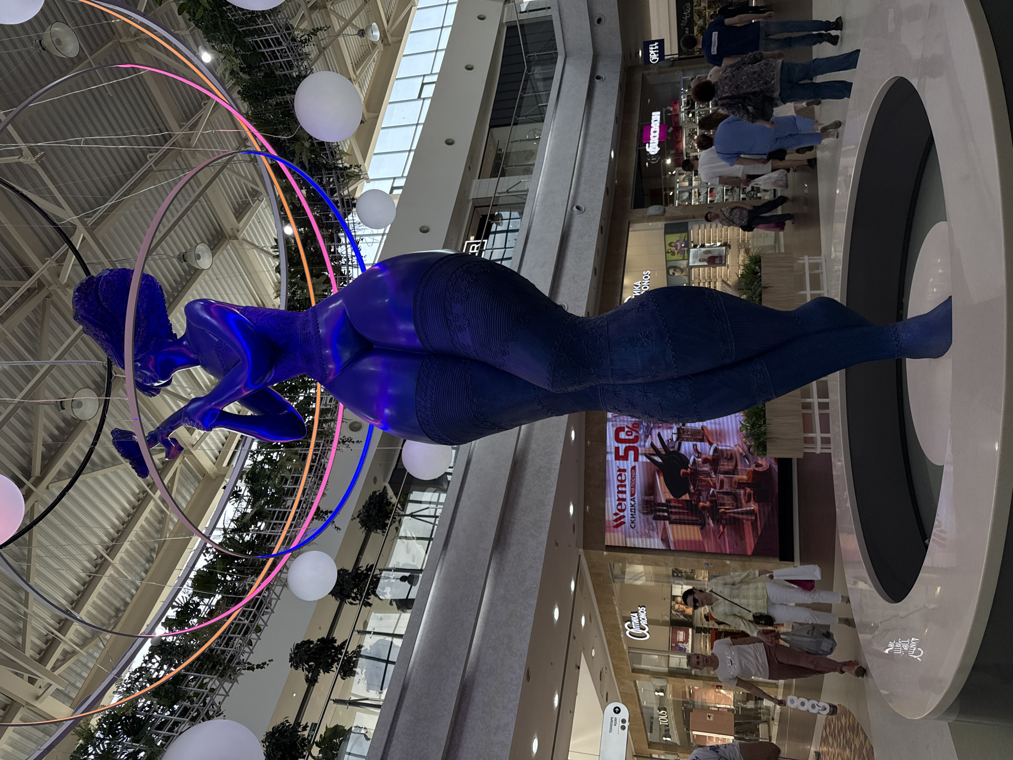 The image is of a large, blue, reclining figure sculpture suspended in a modern shopping mall in Nizhny Novgorod, Russia. The figure is surrounded by illuminated circular bands and illuminated spheres. Overhead, the mall's ceiling features exposed metal beams and a skylight. Below, shoppers are seen walking past store fronts with various signage. The sculpture rests on a white pedestal with a black inset. The image suggests an art installation within a commercial setting.