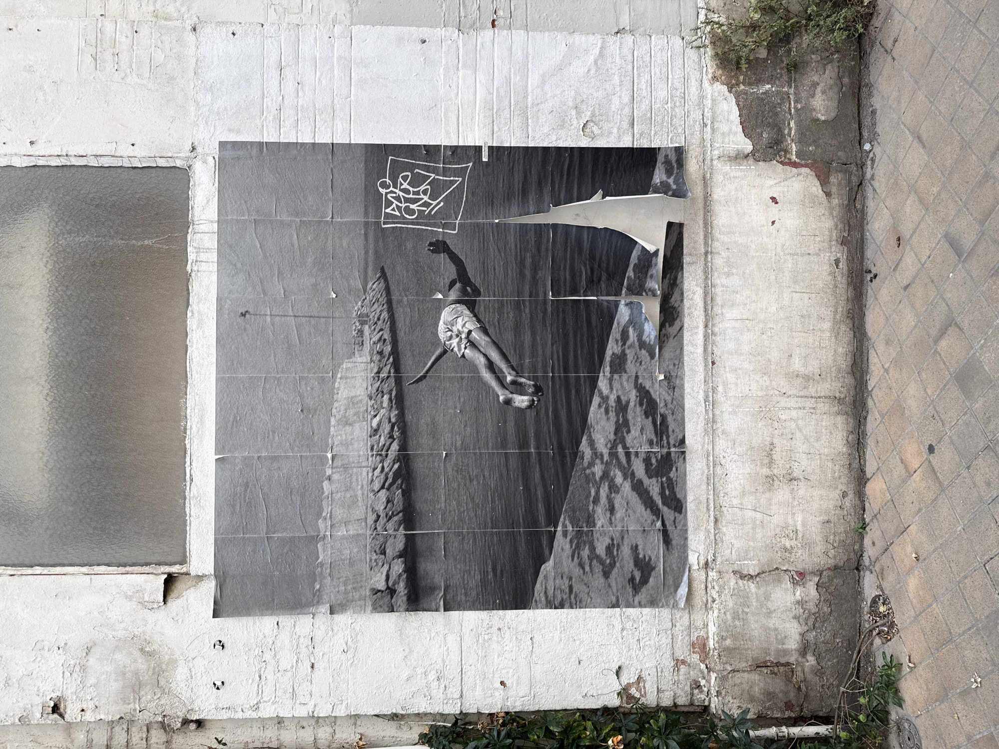 The image shows a large black and white poster pasted on a weathered white brick wall. The poster depicts a person in mid-air, seemingly diving or jumping into water. The individual is wearing patterned shorts and appears to be falling forward. To the right of the person, a white, abstract graffiti tag is drawn within a square outline. The poster is torn and creased in several places, with a large section peeling away at the bottom. The wall itself shows signs of age and decay, with peeling paint and visible brickwork. In the upper part of the image, a frosted glass window is visible. The ground in front of the wall is paved with square tiles. Green vines are growing on the left side of the wall, and some dry leaves and debris are scattered on the ground. The scene suggests an urban environment with a focus on street art and the passage of time. This image is located in Bordeaux, France, specifically on Rue des Ayres.