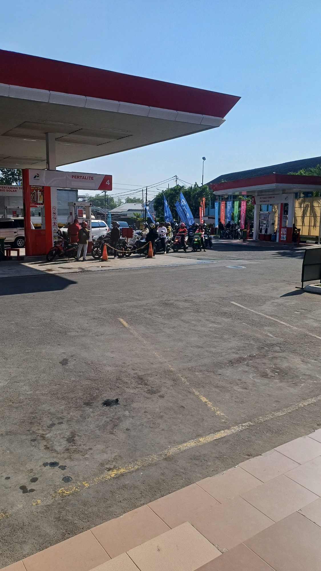 This is a photograph of a Pertamina gas station in Pabelan, Indonesia, on a clear, sunny day. The gas station canopy, painted red and white, extends over multiple gas pumps. Several motorcycles are parked in a line at the pumps, and some individuals are standing near them, possibly refueling or waiting. Blue, red, green, and white flags are displayed prominently in front of the gas station buildings. The ground is paved asphalt, with visible yellow lines marking parking or traffic lanes. In the background, there are buildings and trees, indicating an urban or semi-urban environment.