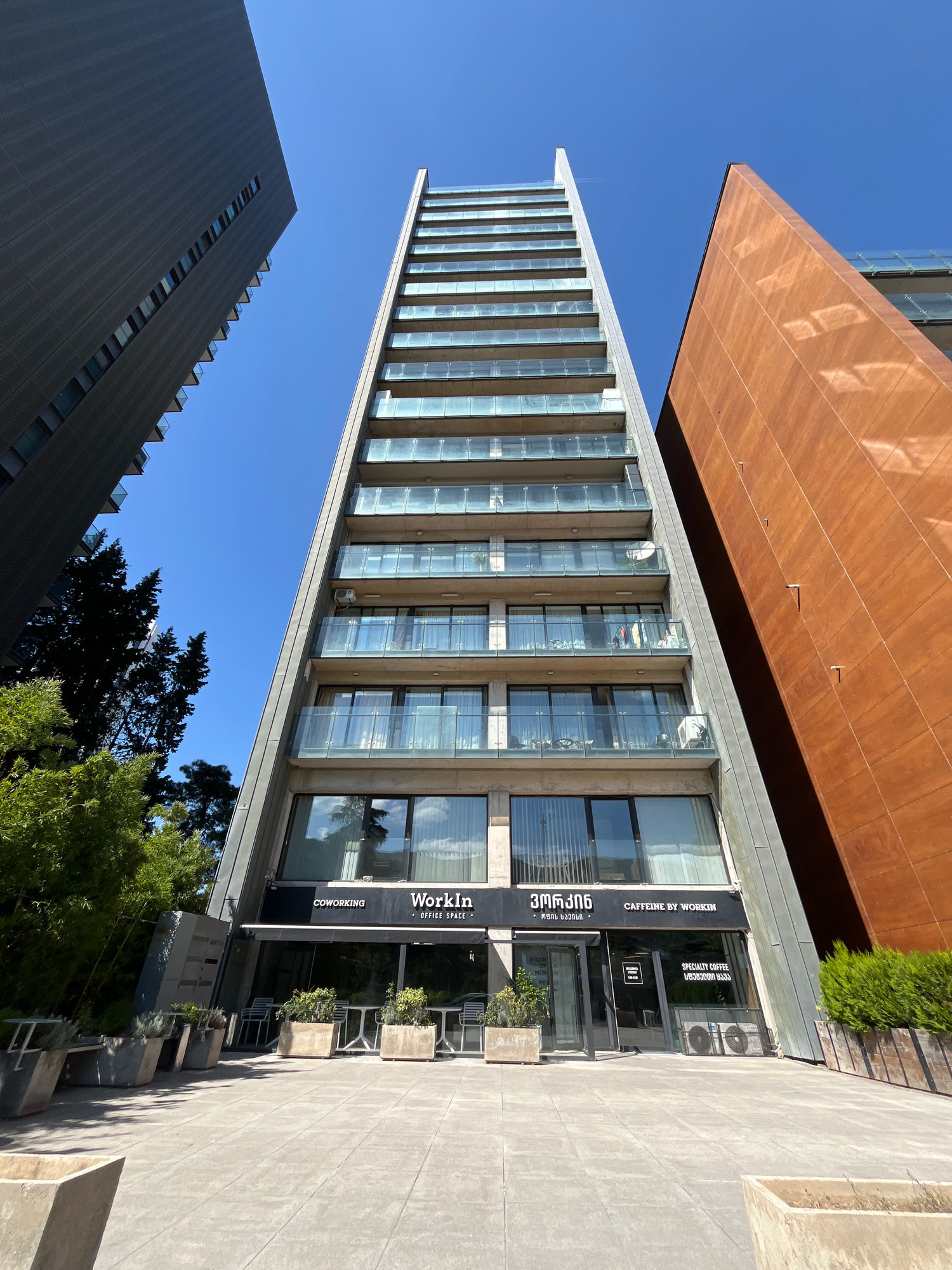 An exterior, ground-level, eye-level, wide shot captures a modern, angular architectural scene on a clear day in Tbilisi, Georgia. The focus is on a tall, narrow building with a stepped facade, featuring multiple balconies with glass railings. Each balcony has some furniture and decorative items visible. The building’s lower level houses a "WorkIn" coworking office space with a cafe called "Caffeine by Workin." The entrance features glass doors and large windows displaying interior seating. Adjacent to this building are two other imposing structures: a dark, textured, tall rectangular building on the left, and a large, reddish-brown, angled building on the right, which appears to be made of metal panels. Green trees and manicured bushes are visible on the left and right sides of the foreground, along with concrete planters. The ground is paved with large, light-colored square tiles. The sky is a vibrant, cloudless blue. No people are discernible in the frame.