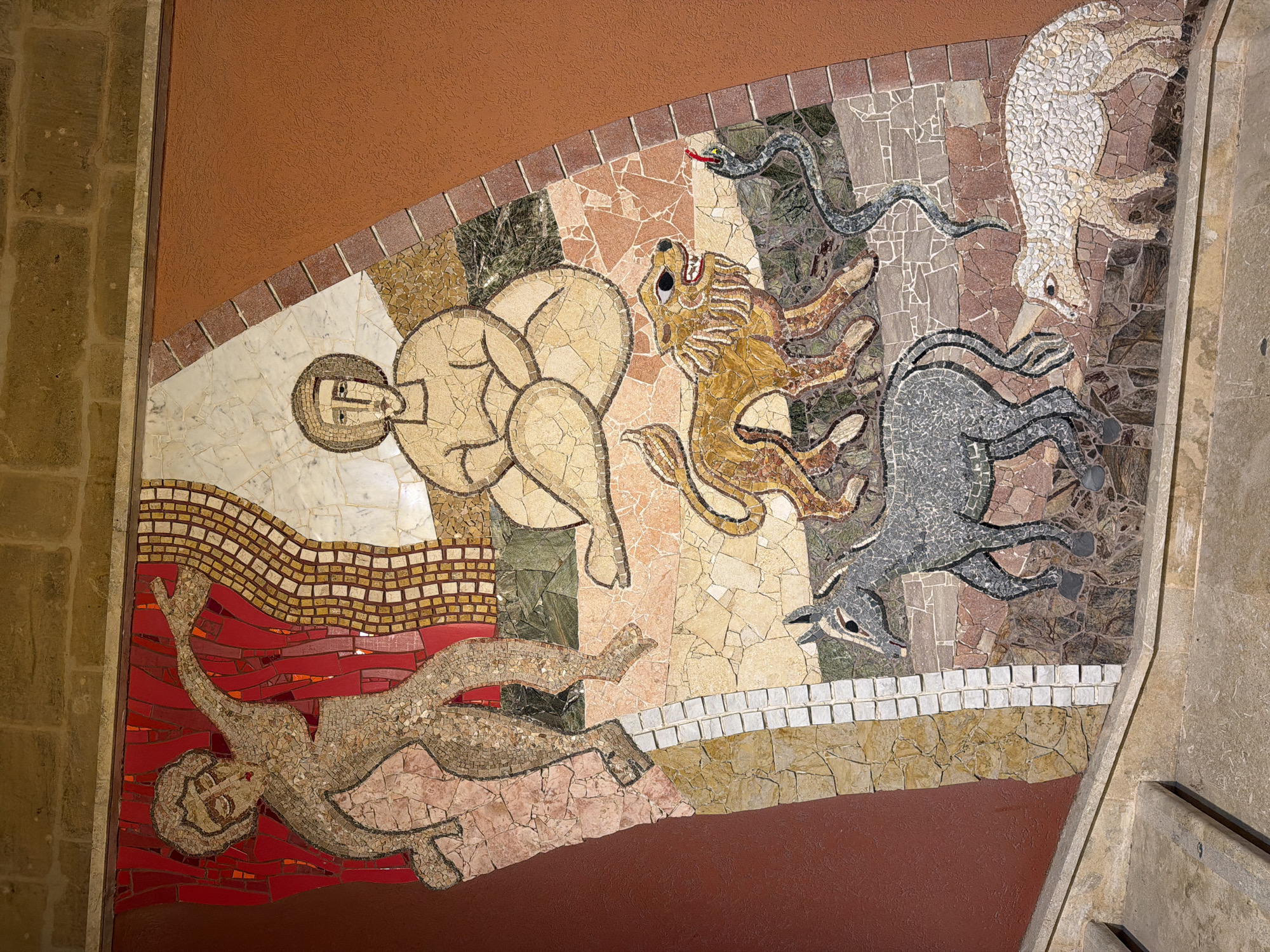 This is a mosaic artwork located on the exterior wall of a building in Victoria, Gozo, Malta. The mosaic depicts a narrative scene composed of various figures and animals rendered in small, colored tiles.
In the upper left, a stylized human figure with outstretched arms is depicted against a red background, with flowing red and gold mosaic patterns behind it. To the right of this figure, another human figure sits cross-legged, with a halo-like circular mosaic around its head, against a white and beige background.
Below these figures, a sequence of animals is shown. A lion with a mane and open mouth is depicted in the center, looking towards a dark grey snake coiled to its right. In front of the lion, a grey donkey or mule is shown in profile, facing left. At the bottom right of the mosaic, a white lamb or sheep with a distinct horn is walking towards the left.
The mosaic is set against a reddish-brown textured wall, with a section of beige tiled steps visible in the lower left of the frame, indicating it is likely part of an outdoor staircase or entrance. The mosaic itself is bordered by sections of stone and brickwork, suggesting it is an integrated architectural feature. The overall style of the mosaic appears somewhat primitive or folk-art inspired, with simplified forms and bold outlines.