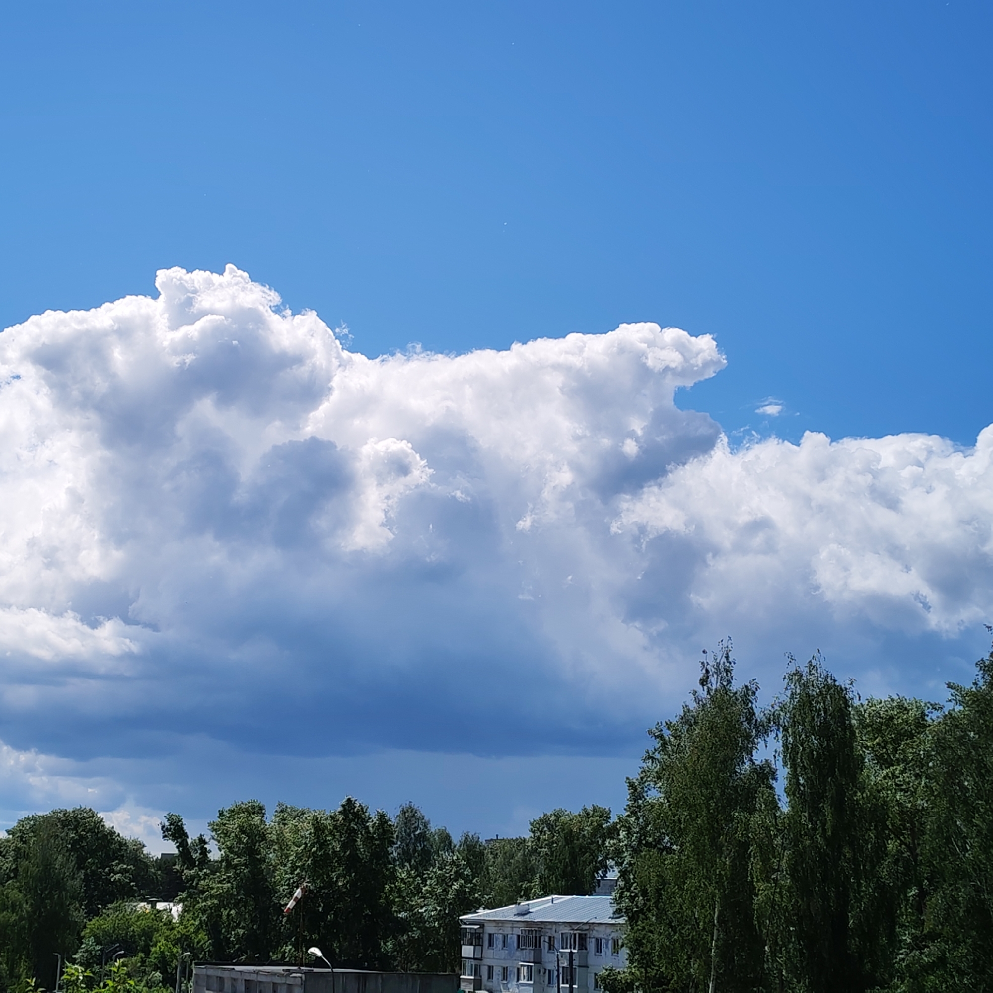 The image shows a daytime view in Chaikovsky, Russia. A large, puffy cumulonimbus cloud dominates the sky above the tree line. Below the cloud, a building with white walls and a gray roof is visible among the green trees. A light post is visible to the left of the building. The sky is a clear blue in the upper portion of the frame.
