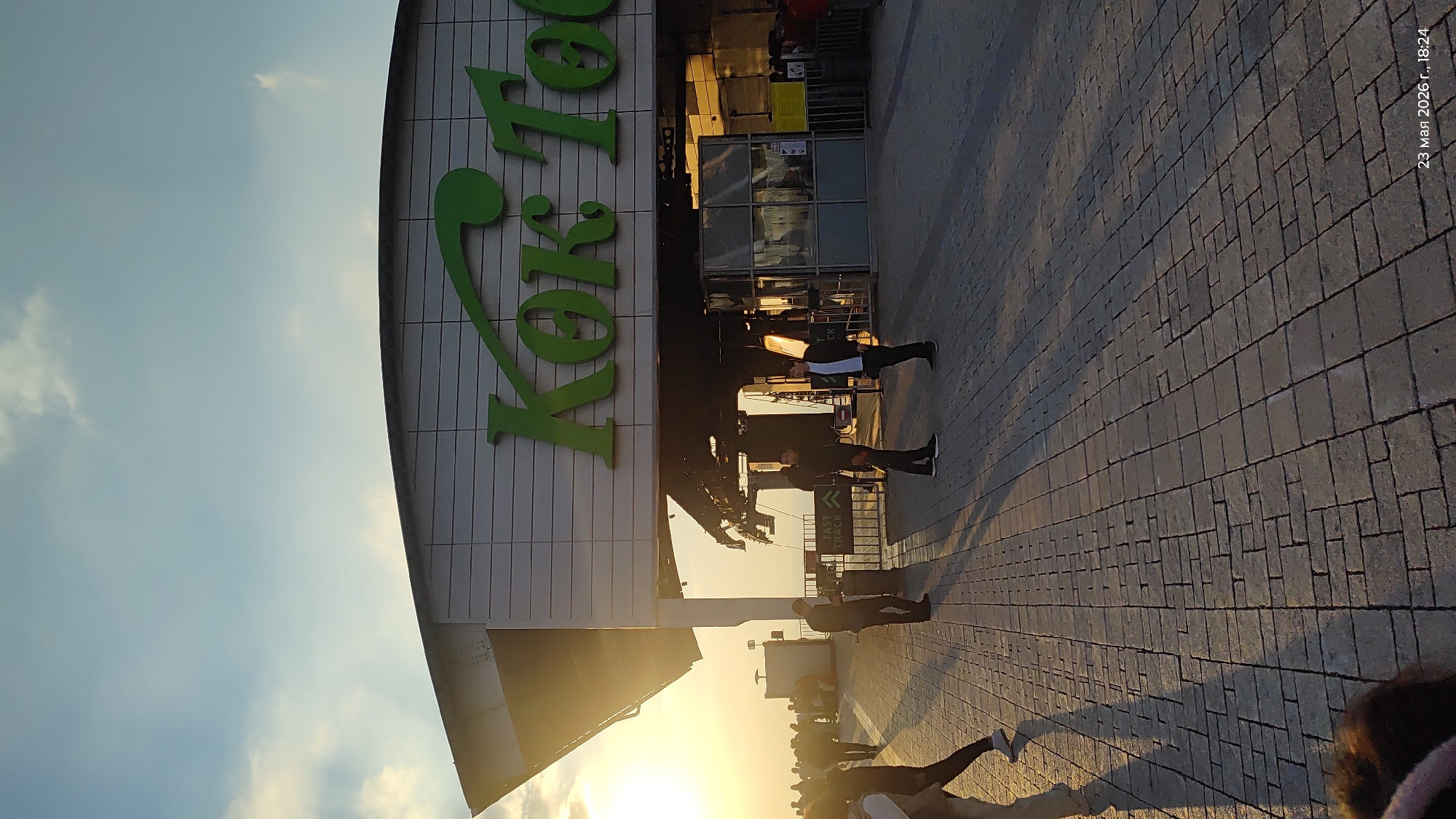Here is a description of the image:
The image shows the exterior of a "Koktobe" supermarket or shopping center, judging by the large green sign. People are seen walking on the paved walkway in front of the building. The photo appears to be taken during sunset, with the sun shining brightly. The image was taken on March 23, 2026, at 18:24. The location is most likely Almaty, Kazakhstan, given the name of the building.