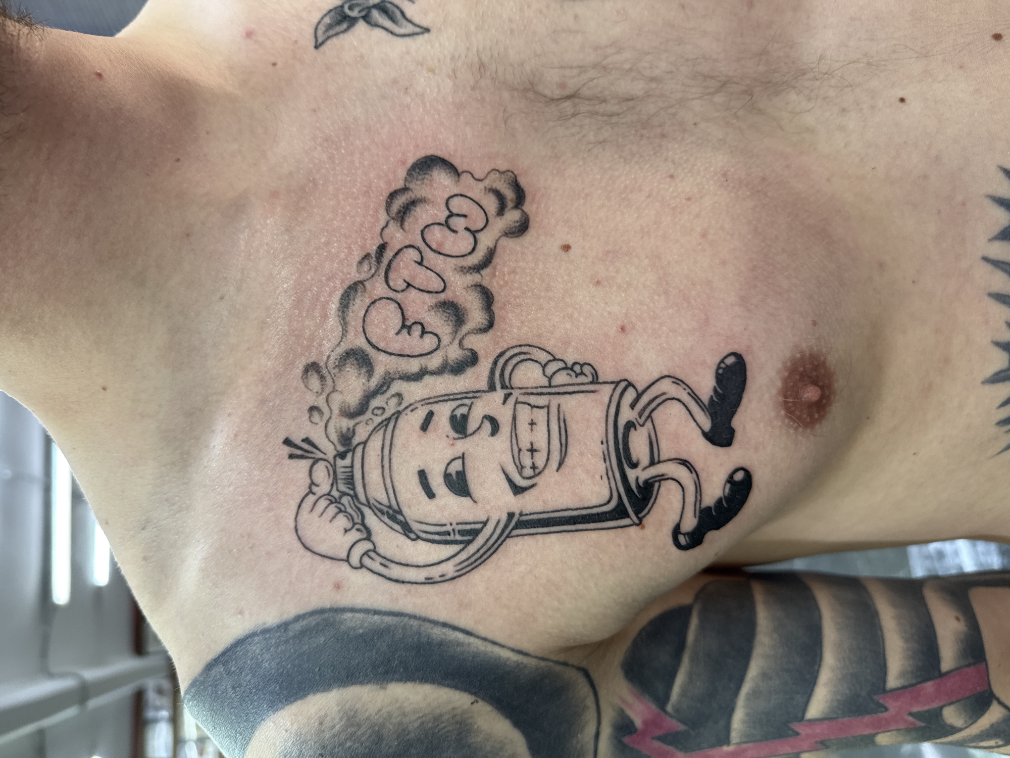 The image captures a close-up of a person's upper torso, showcasing a prominent black and white tattoo on their chest. The individual has a light skin tone with some visible chest hair. The central tattoo features an anthropomorphic spray paint can character, depicted with a wide, happy grin, eyes, a nose, and small arms and legs wearing black shoes. From the nozzle of the spray can, a stylized cloud of smoke or gas billows upwards, containing the letters "CBCO" in a rounded, bubble-like font.
The skin around the tattoo shows slight redness, suggesting it may be a recent or fresh piece of body art. Other tattoos are partially visible on the person's body; a small, dark leaf design is seen near the collarbone on the right, and a large black and white striped pattern with a striking pink lightning bolt is present on the right arm, extending towards the chest. A nipple is visible on the right side of the chest, below the main tattoo. The background is out of focus, indicating an indoor setting, possibly a tattoo studio, with vertical elements suggesting windows or blinds. The overall scene is a personal display of body art, located in Moscow, Russia.