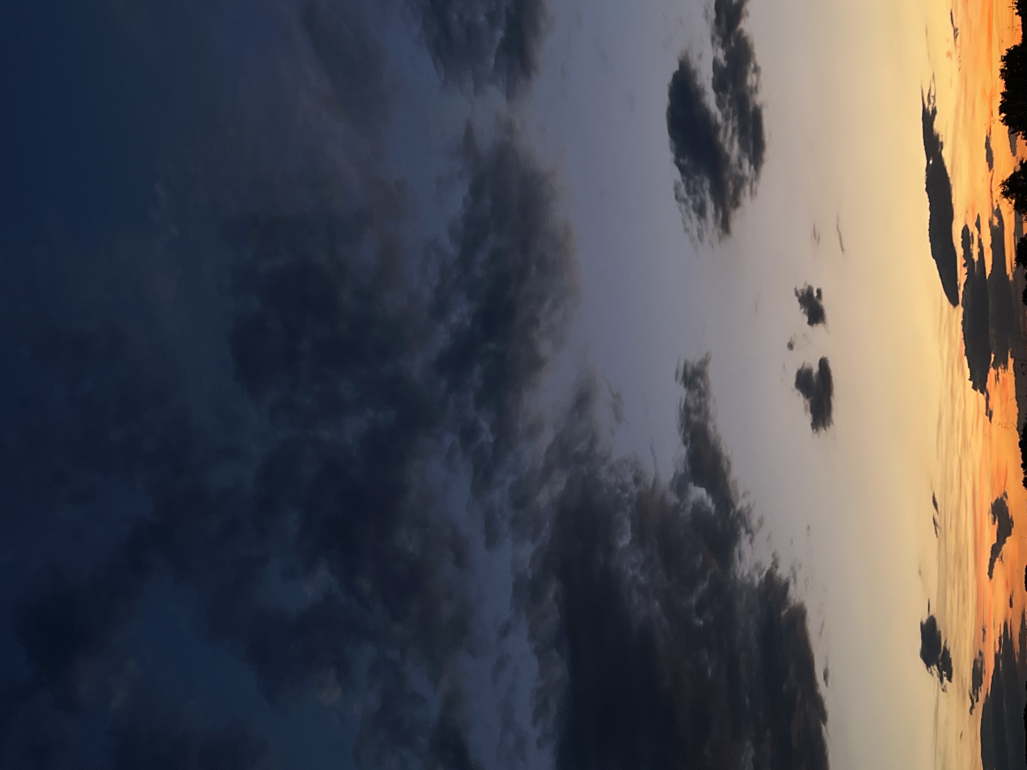The image shows a sunset sky over Plovdiv, Bulgaria. The sky transitions from dark blue at the top to hues of purple, orange, and yellow near the horizon. Scattered dark clouds are present throughout the sky, with some appearing more densely packed at the top and others more wispy and spread out towards the bottom. The very bottom of the image shows the silhouette of trees and possibly some low-lying structures, suggesting a natural or semi-rural setting within or on the outskirts of the city. There are no people, animals, or distinct man-made objects visible, beyond the faint silhouette at the horizon. The overall impression is of a serene, natural landscape at dusk.