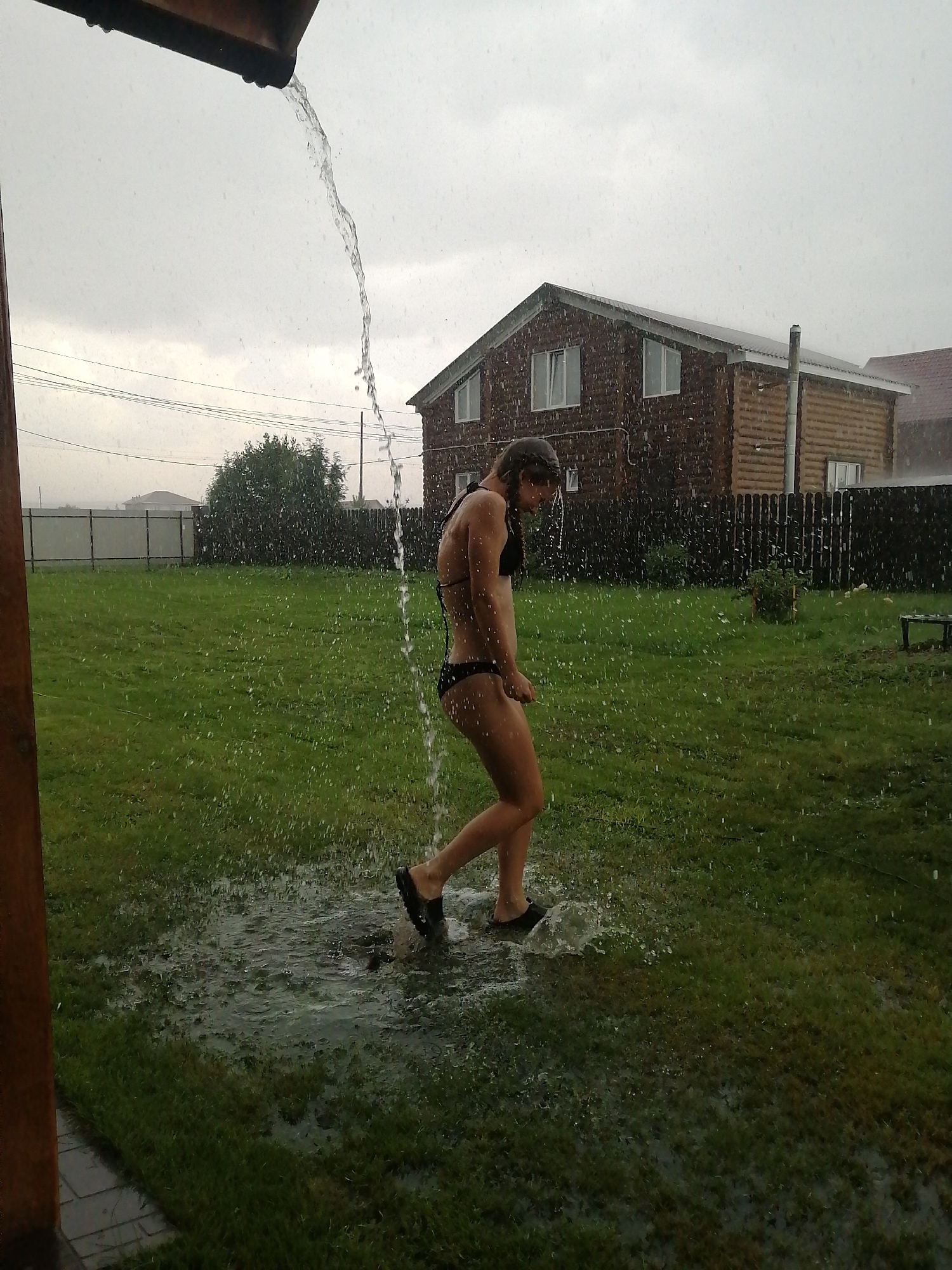 In Yemelyanovo, Russia, a person in a black bikini stands in a grassy yard, receiving a stream of water from an overhead gutter. The water cascades down, creating puddles on the lawn. In the background, a two-story wooden house with visible windows and a fence are situated. The sky is overcast, and raindrops are evident throughout the image, suggesting it is raining. The scene captures a moment of outdoor activity amidst inclement weather.